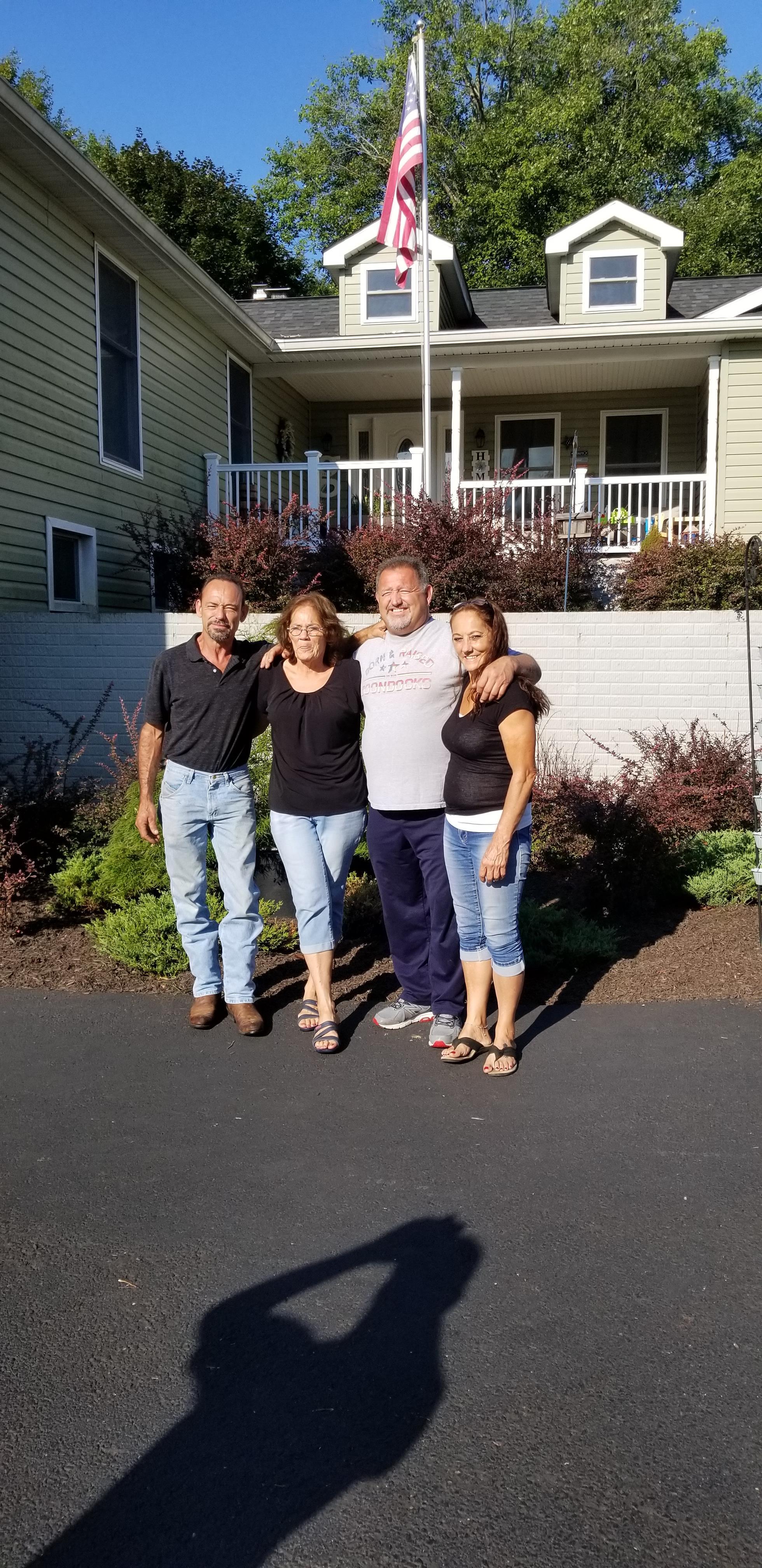 Four people stand close together in front of a house. It is sunny and warm outside.