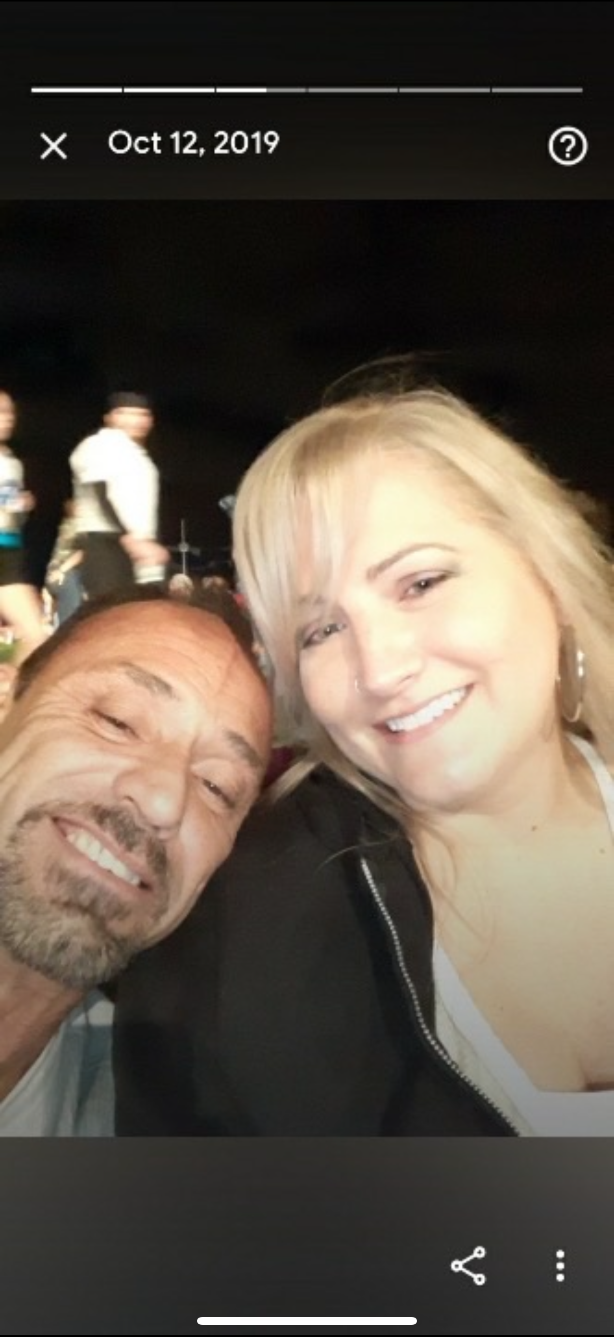A couple smiles widely while sitting close together at a busy gathering in the evening.