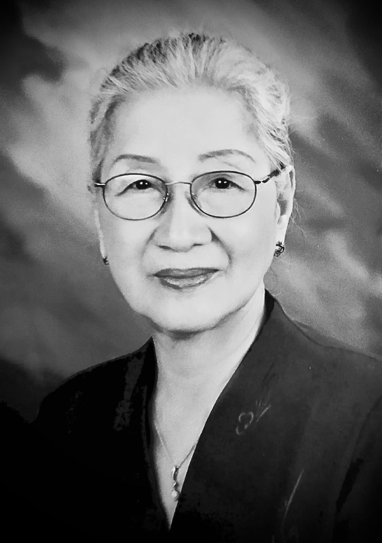 An elderly woman stands and looks at the camera wearing glasses and a dark outfit.