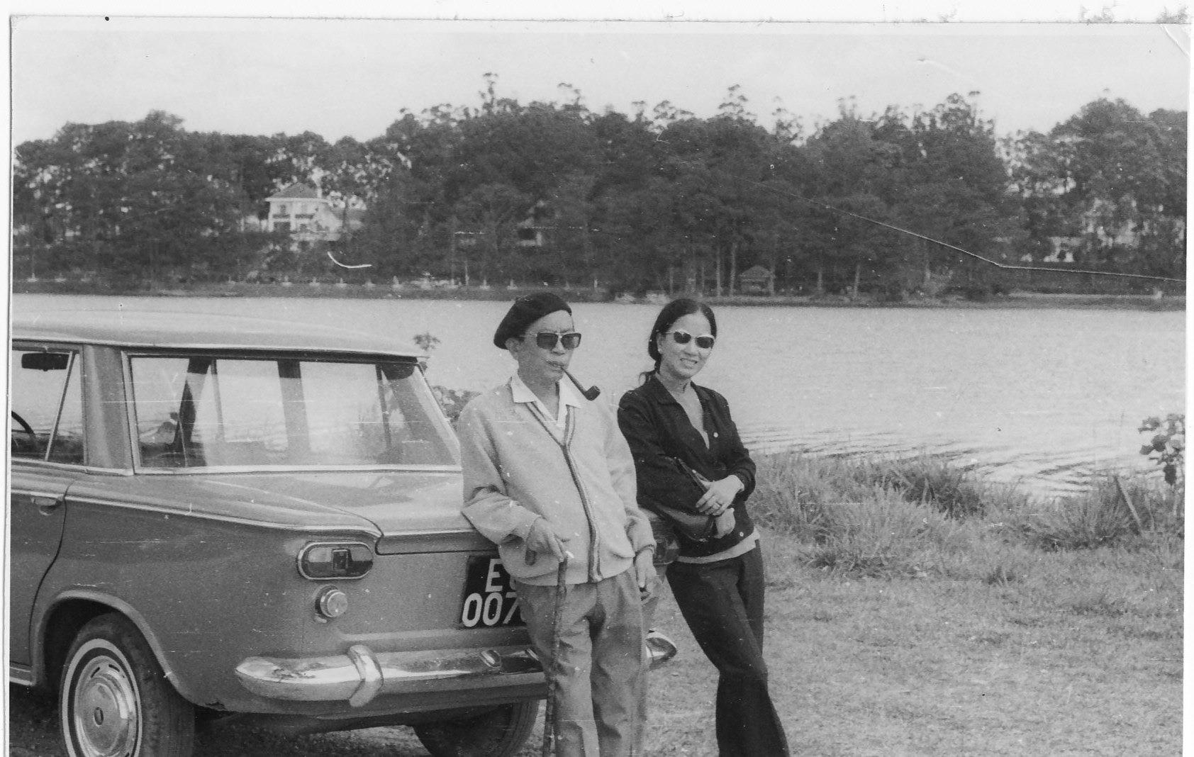 Two individuals pose next to a parked car beside a lake surrounded by trees during daytime.