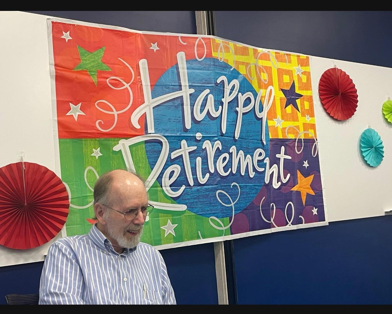 A man sits smiling in front of a large retirement banner during a gathering in an office.