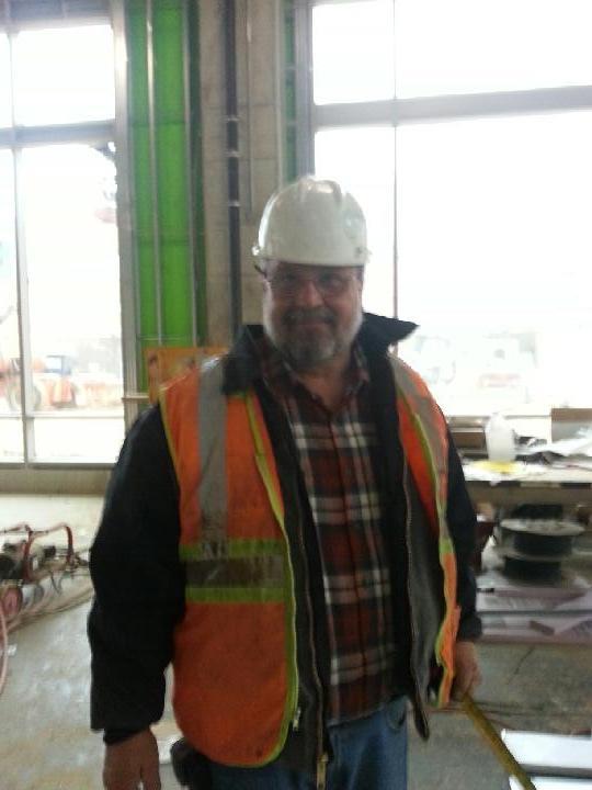 A construction worker stands at a building site wearing a safety vest and hard hat.