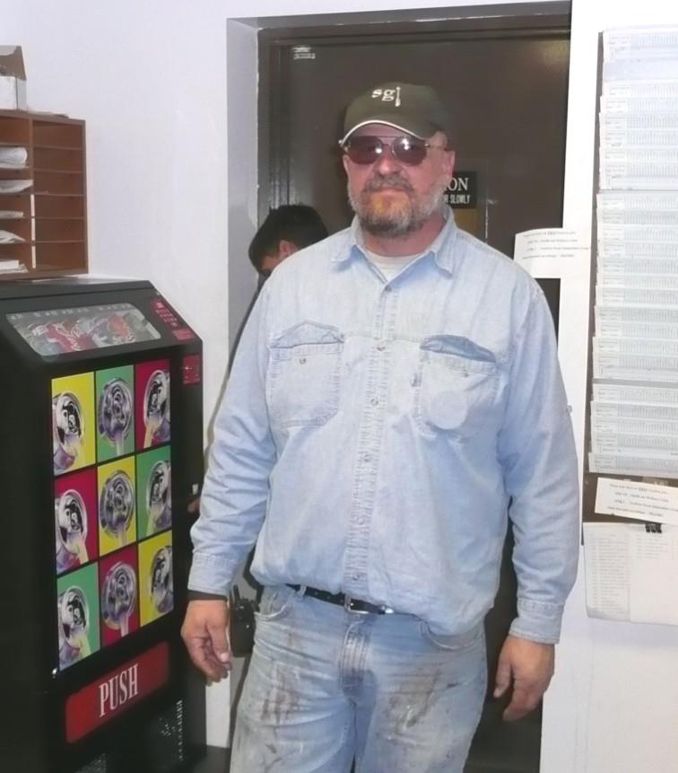 A man in denim clothing stands by a vending machine in a hallway. He looks relaxed and casual.