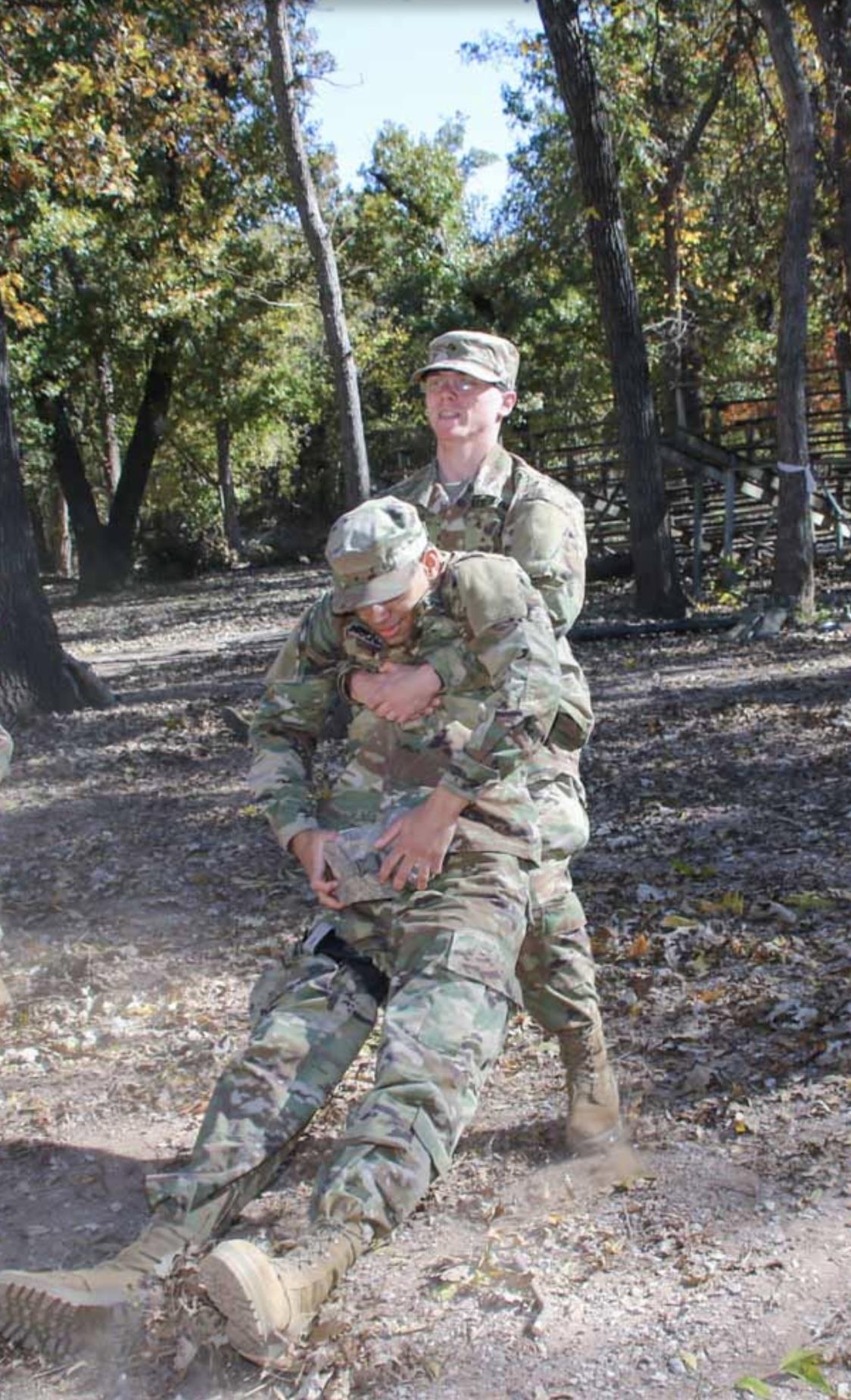 Two soldiers engage in a training drill in a wooded area during daylight hours.
