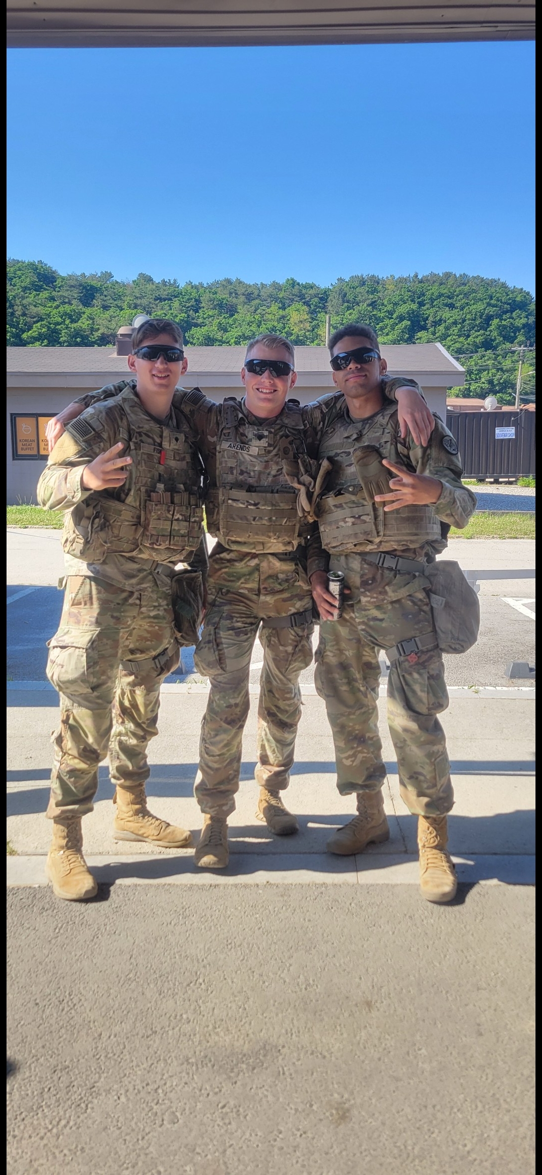 Three soldiers stand together outside a building wearing military gear and smiling proudly.