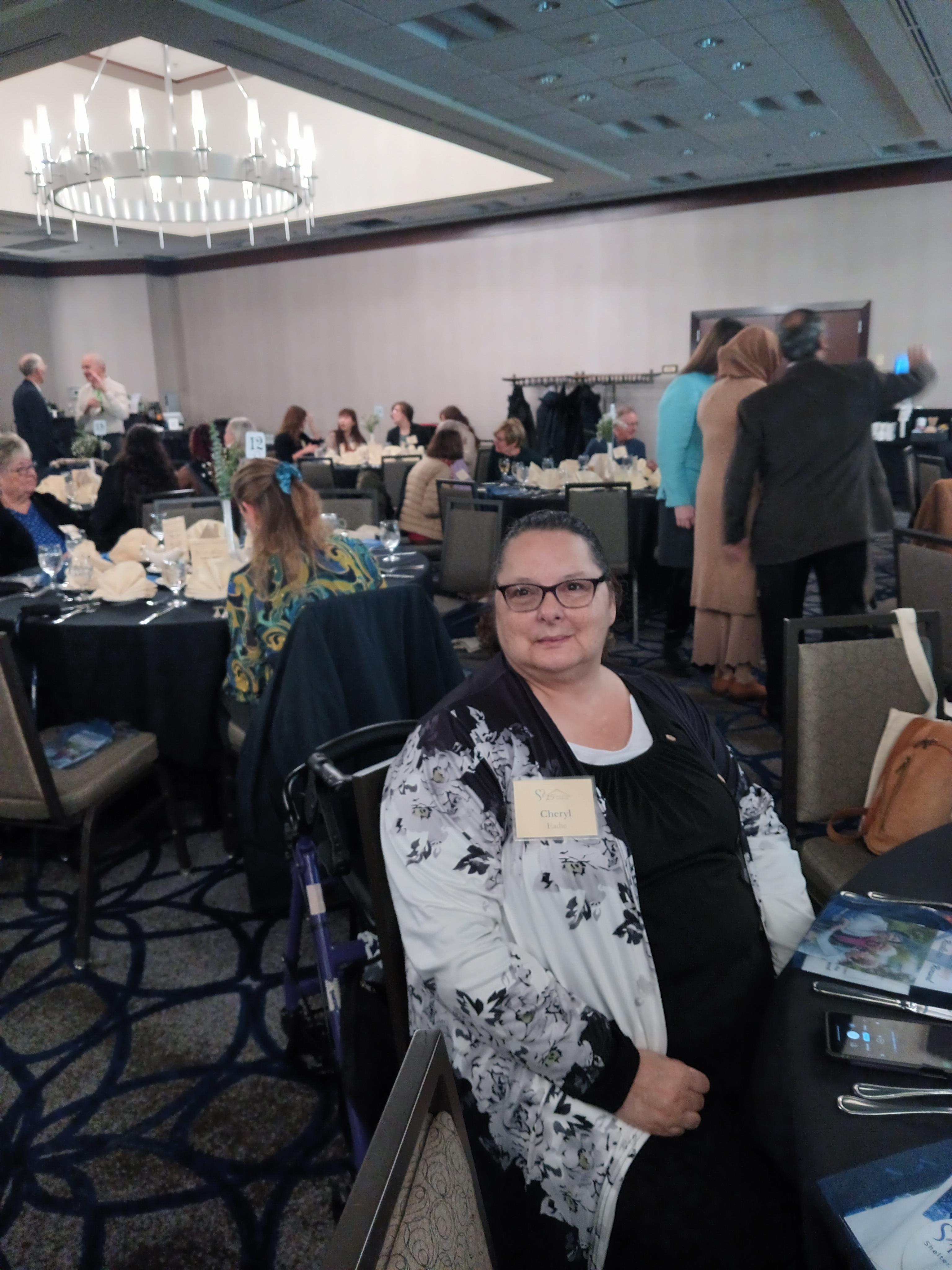 A woman sits at a table while people gather around during a busy event inside a large venue.