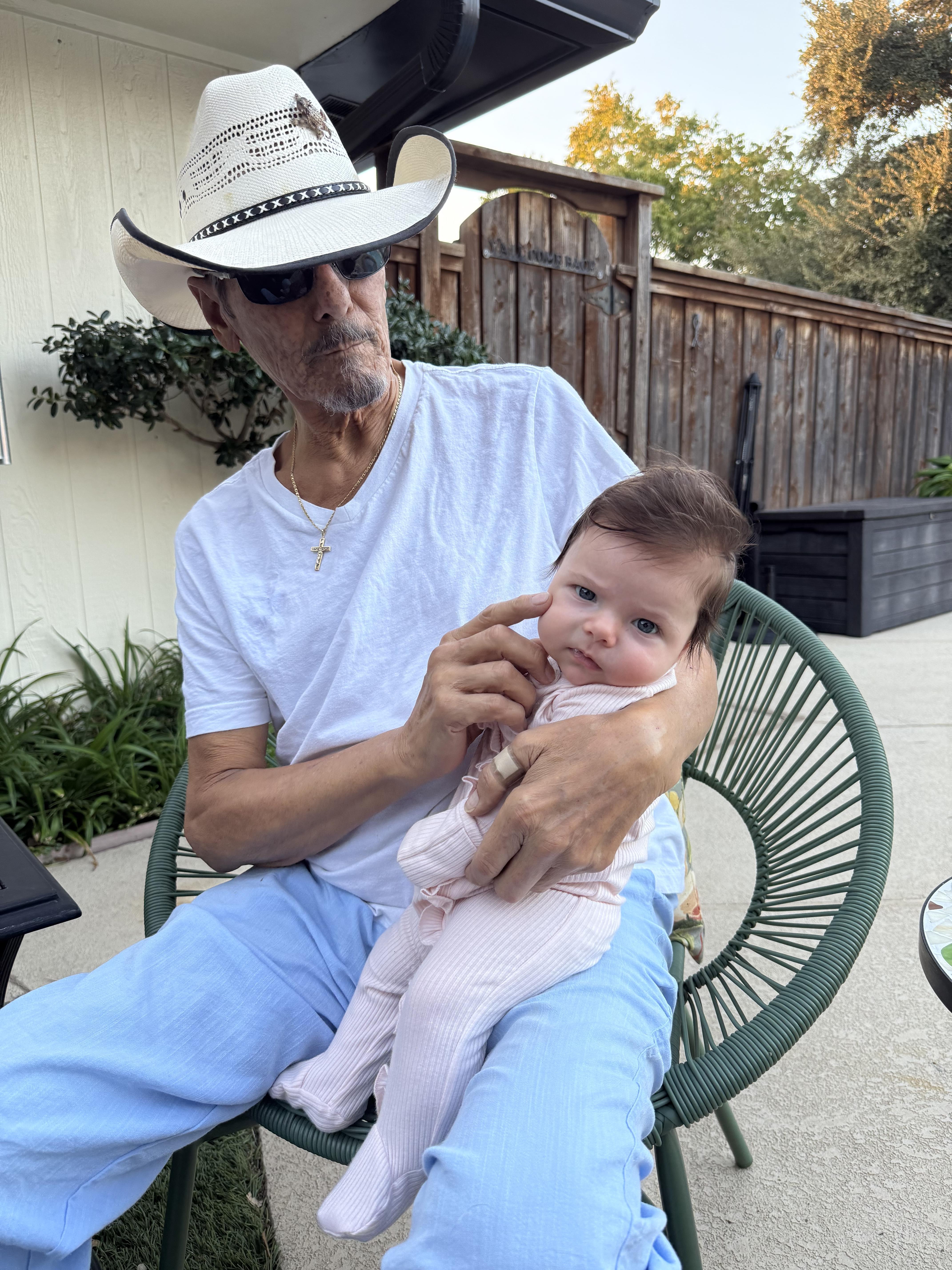 A man sits in a chair with a baby in his lap, both smiling while enjoying the outdoors.