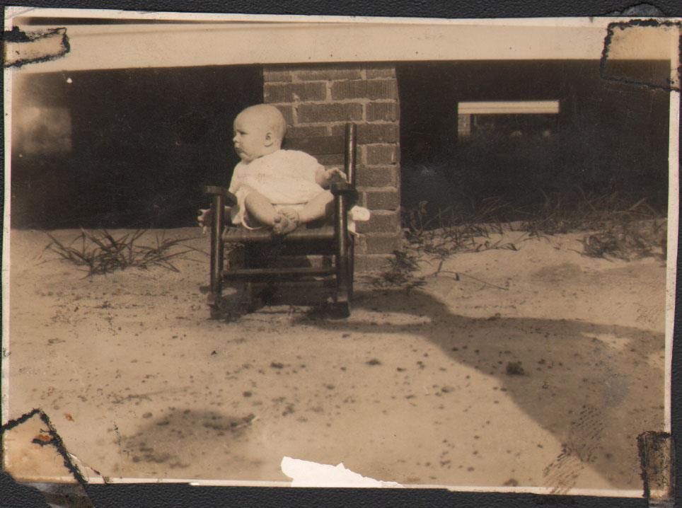 A baby relaxes in a small chair by a brick structure with dirt and grass around.
