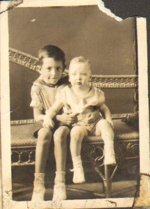 Two children pose together on a couch in a vintage room. One child sits beside the other.