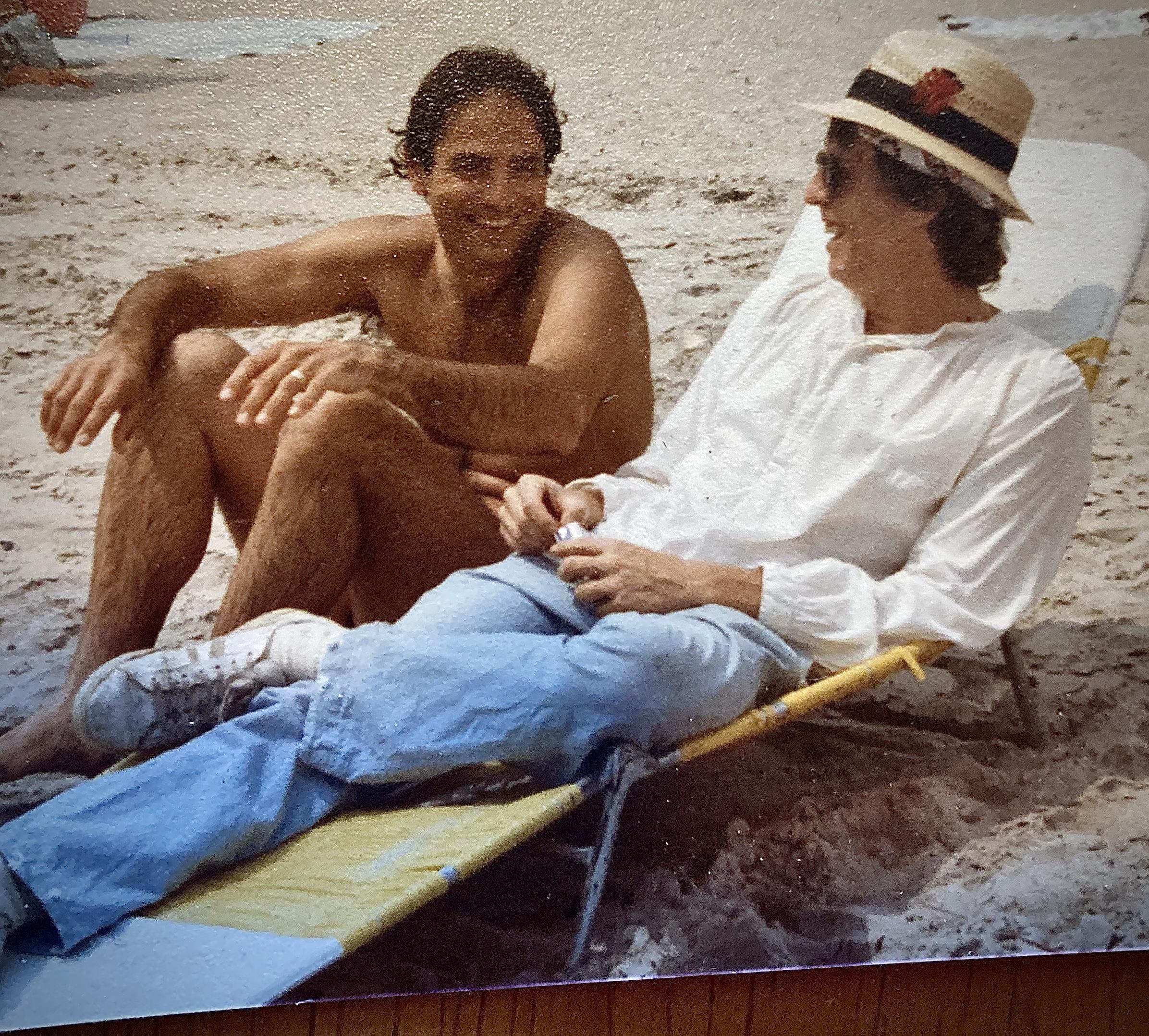 Two men sit on the sand at the beach. They are talking and laughing while enjoying the warm sun.