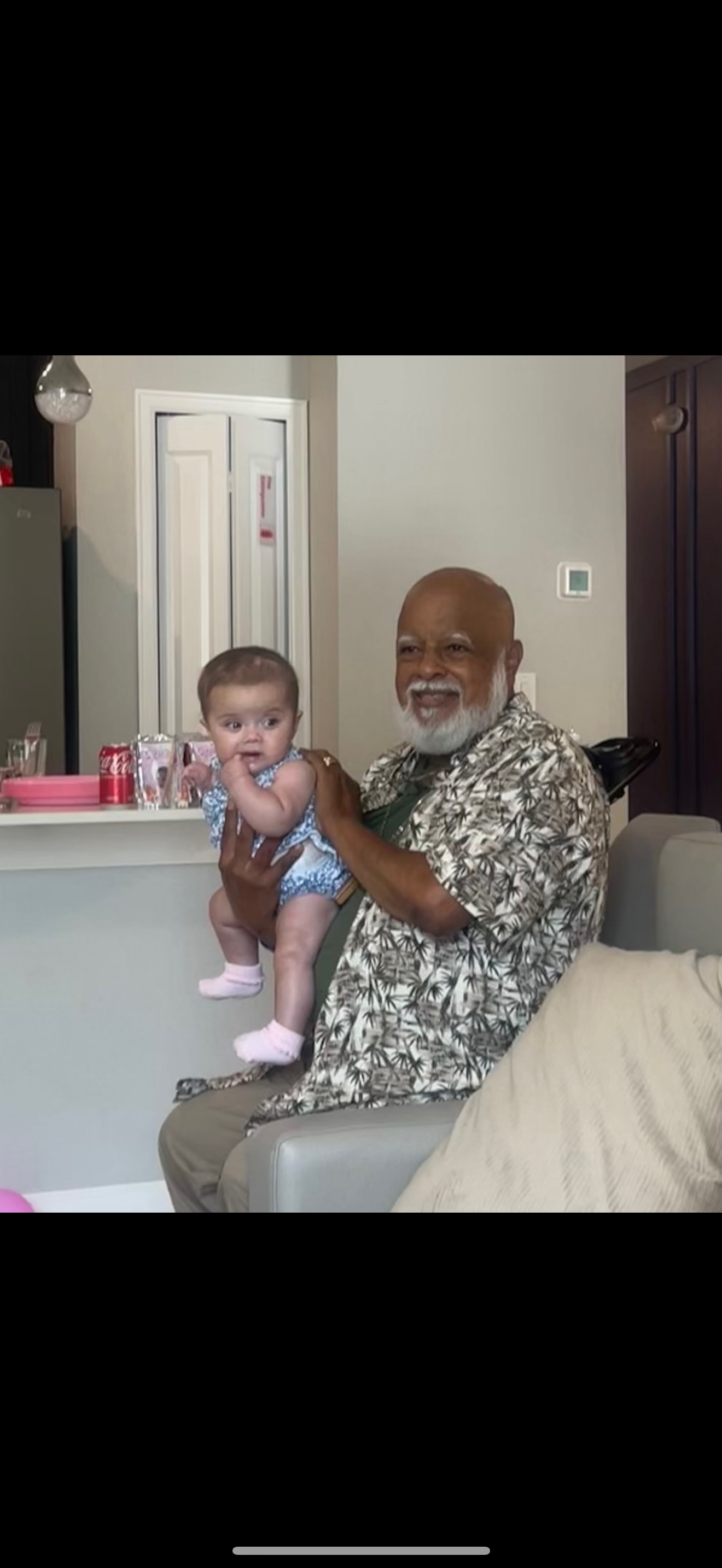 An elderly man sits on a couch holding a baby girl in a living room during a family event.