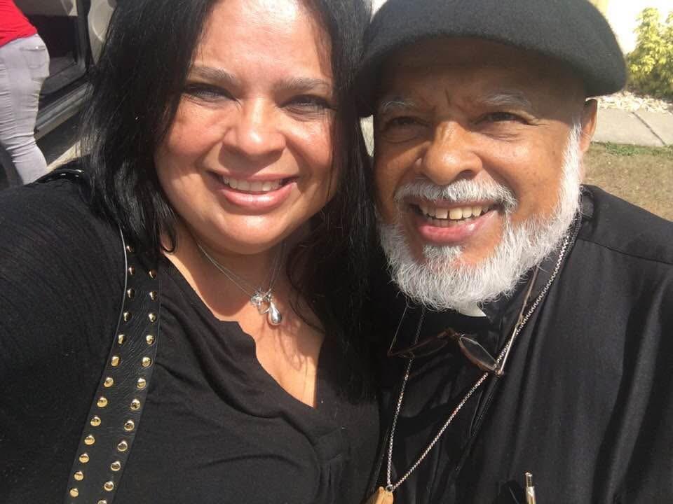Two adults stand together outside and smile at the camera on a bright day.