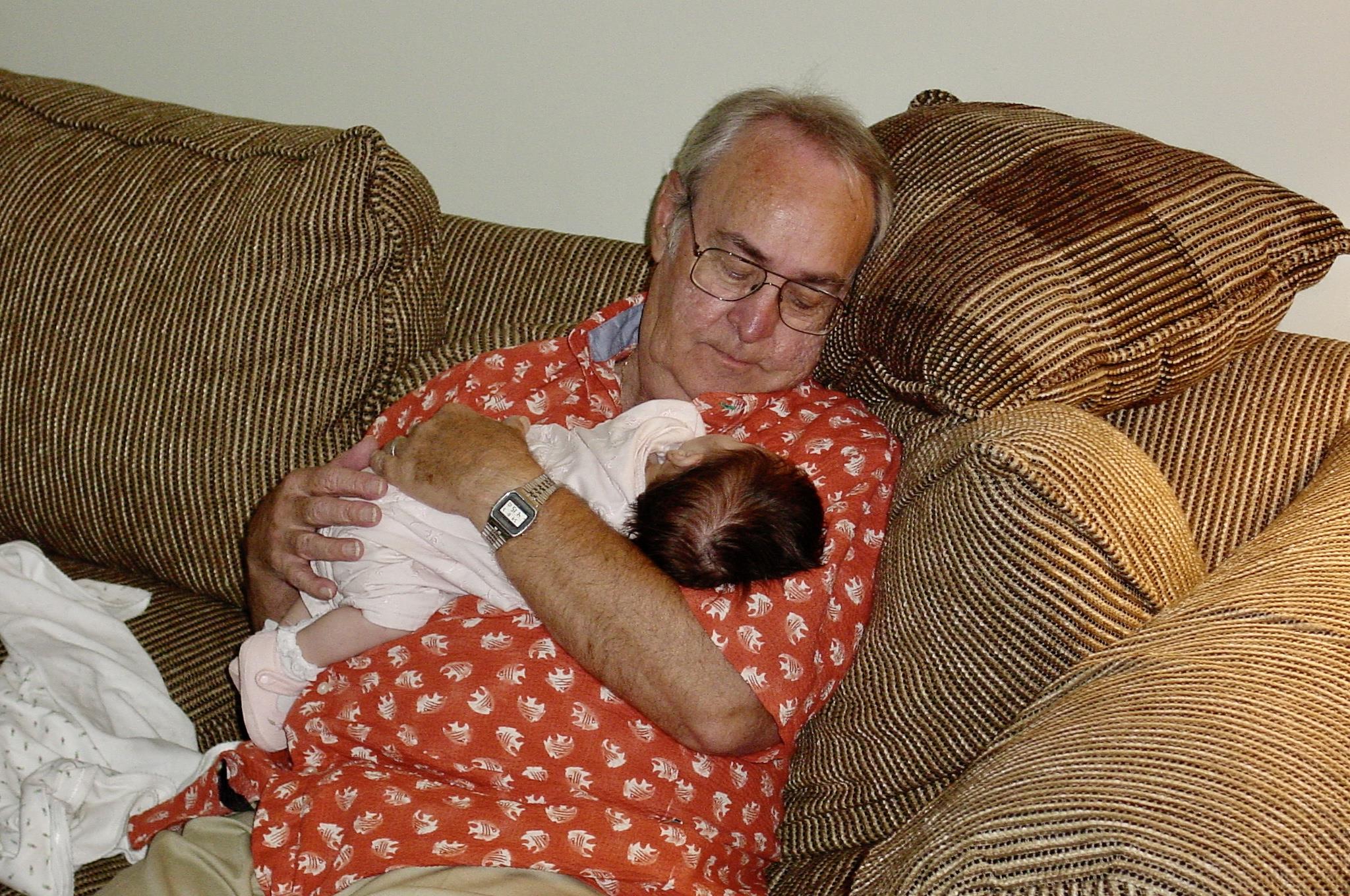 A grandfather sits on a couch and holds a baby in his arms. They both look relaxed and quiet.