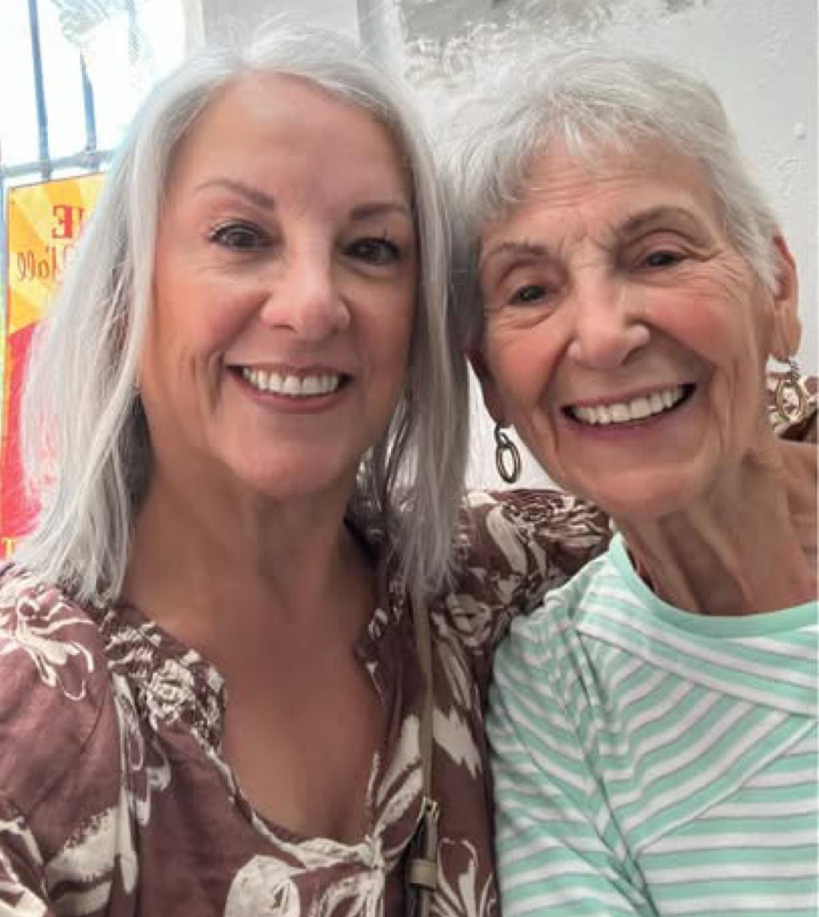 Two women pose happily for a picture in a brightly lit room with light colors and cheerful smiles.