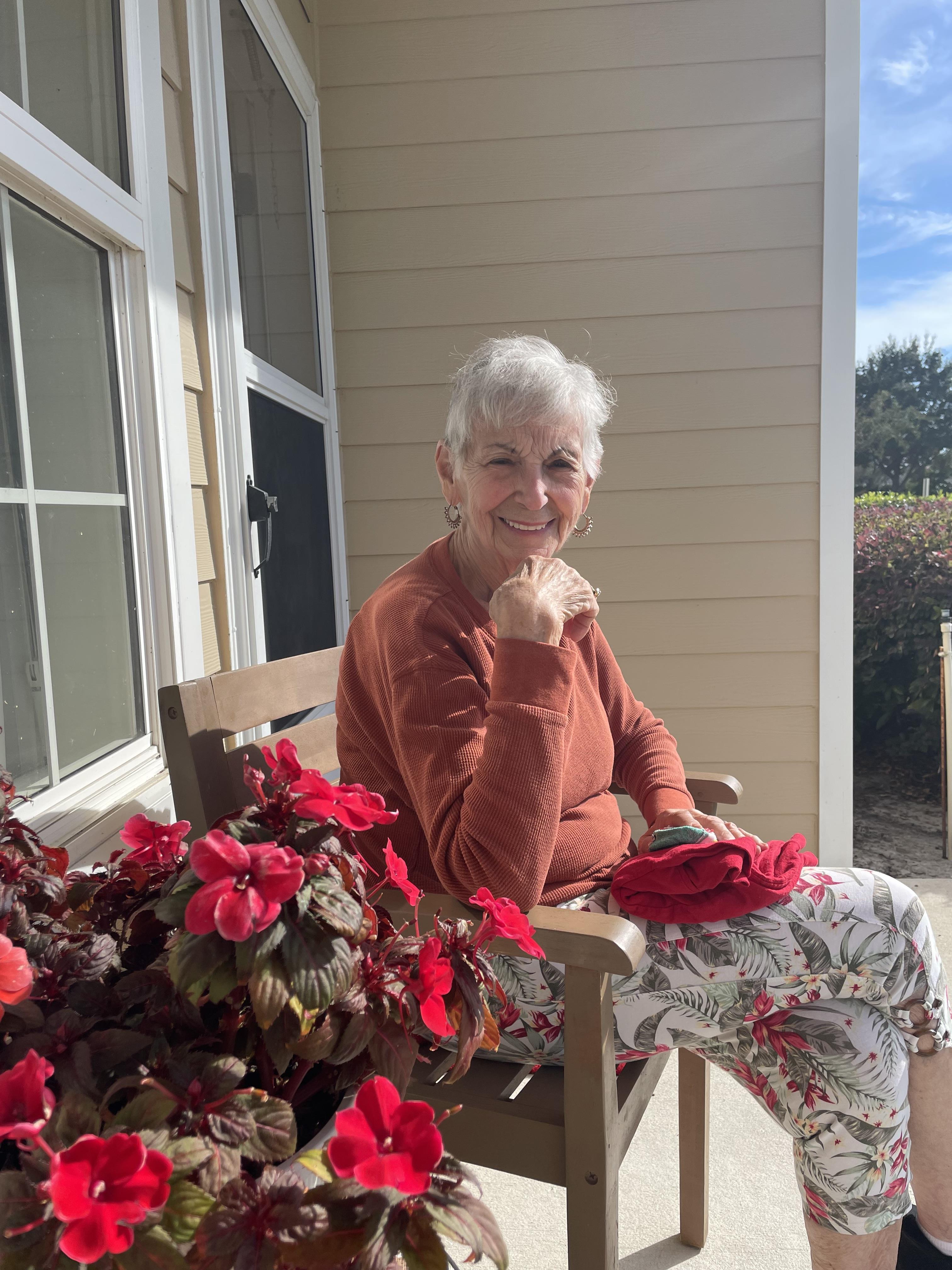 Older woman sits on a porch with flowers and smiles while holding a piece of cloth.
