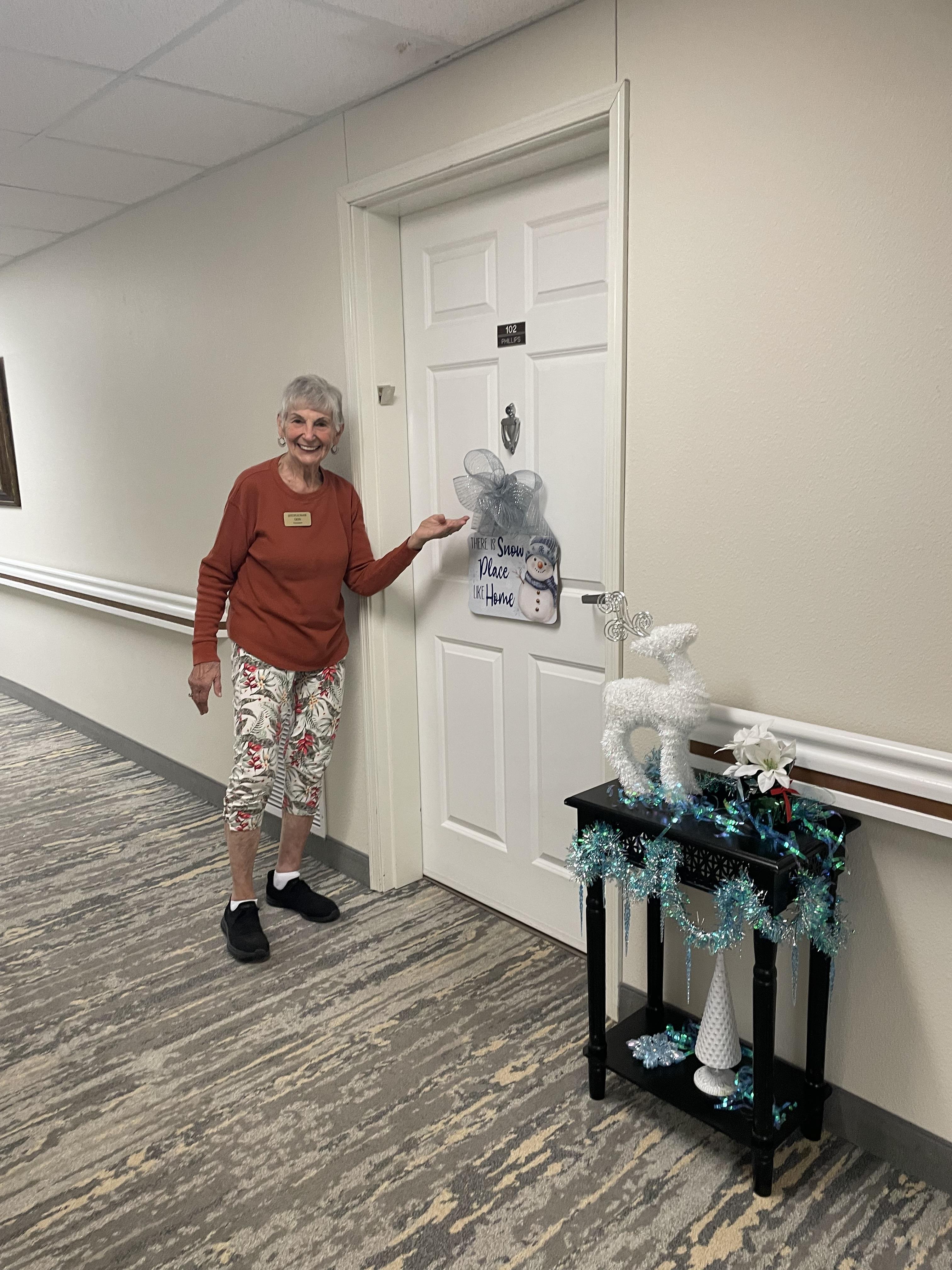 A senior resident smiles in front of a decorated door in a care facility hallway.
