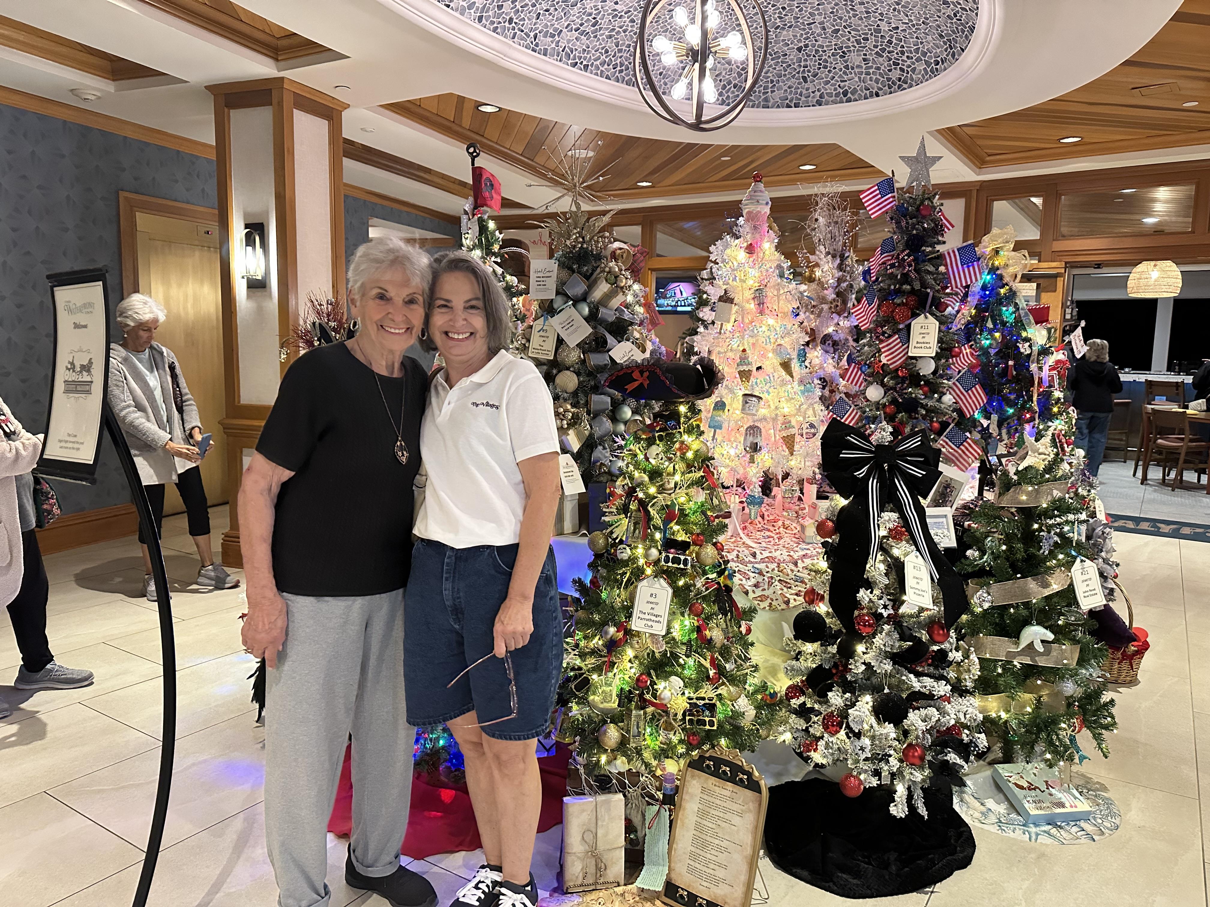 Two women smile and stand next to beautiful Christmas trees in a busy area during a holiday event.