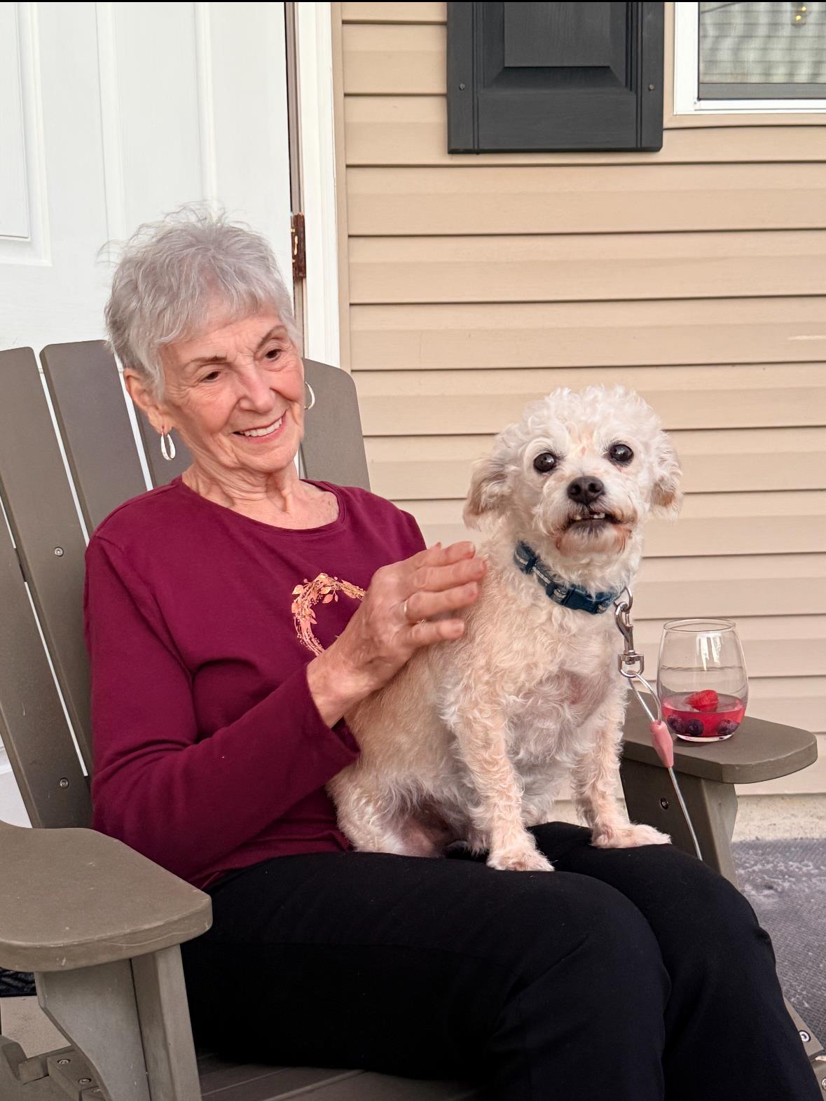 A woman smiles as she pets a small dog on her lap while sitting outside at home.