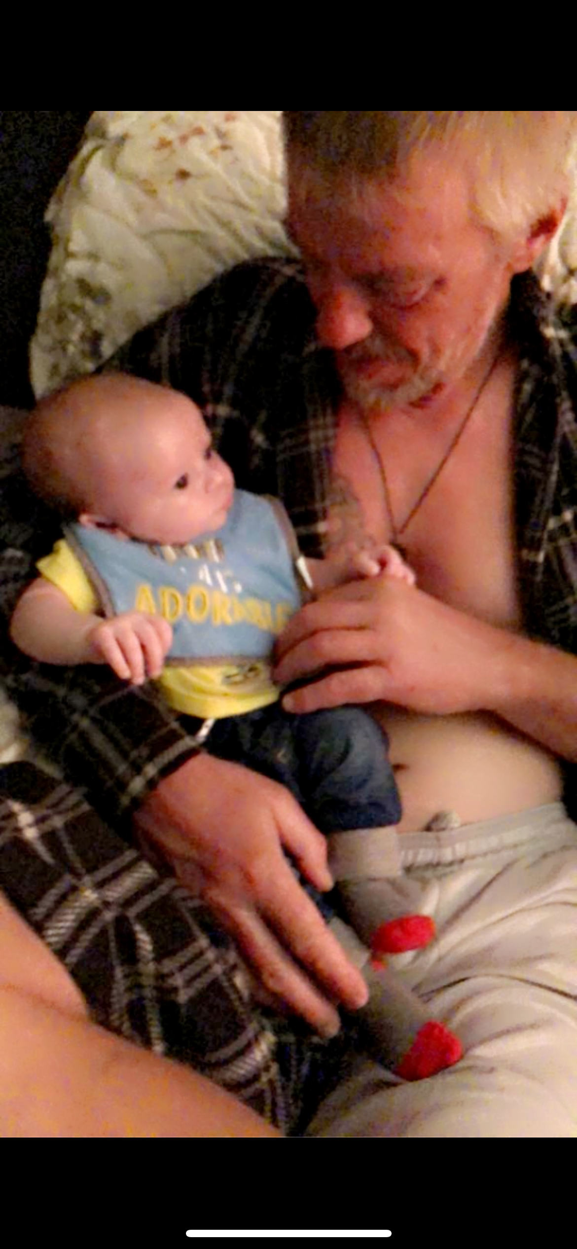 A grandfather is sitting and holding a baby wearing a bib in a relaxed moment at home.
