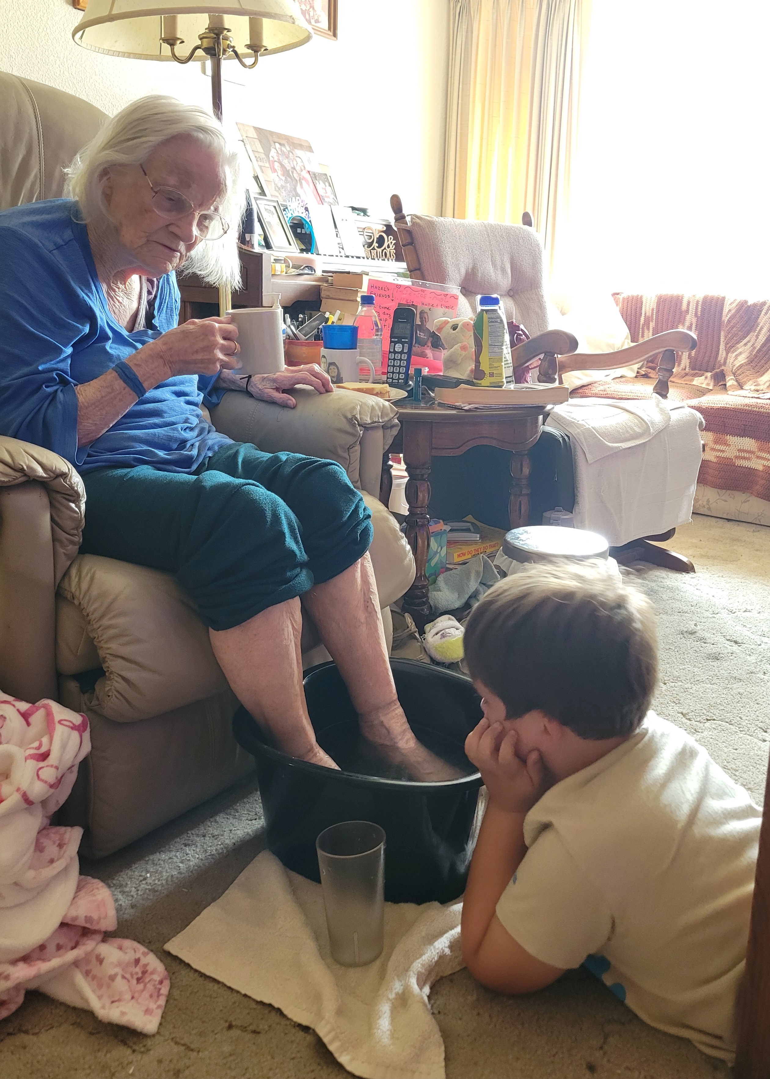 An elderly person sits in a chair with a cup, while a child observes closely as feet soak.