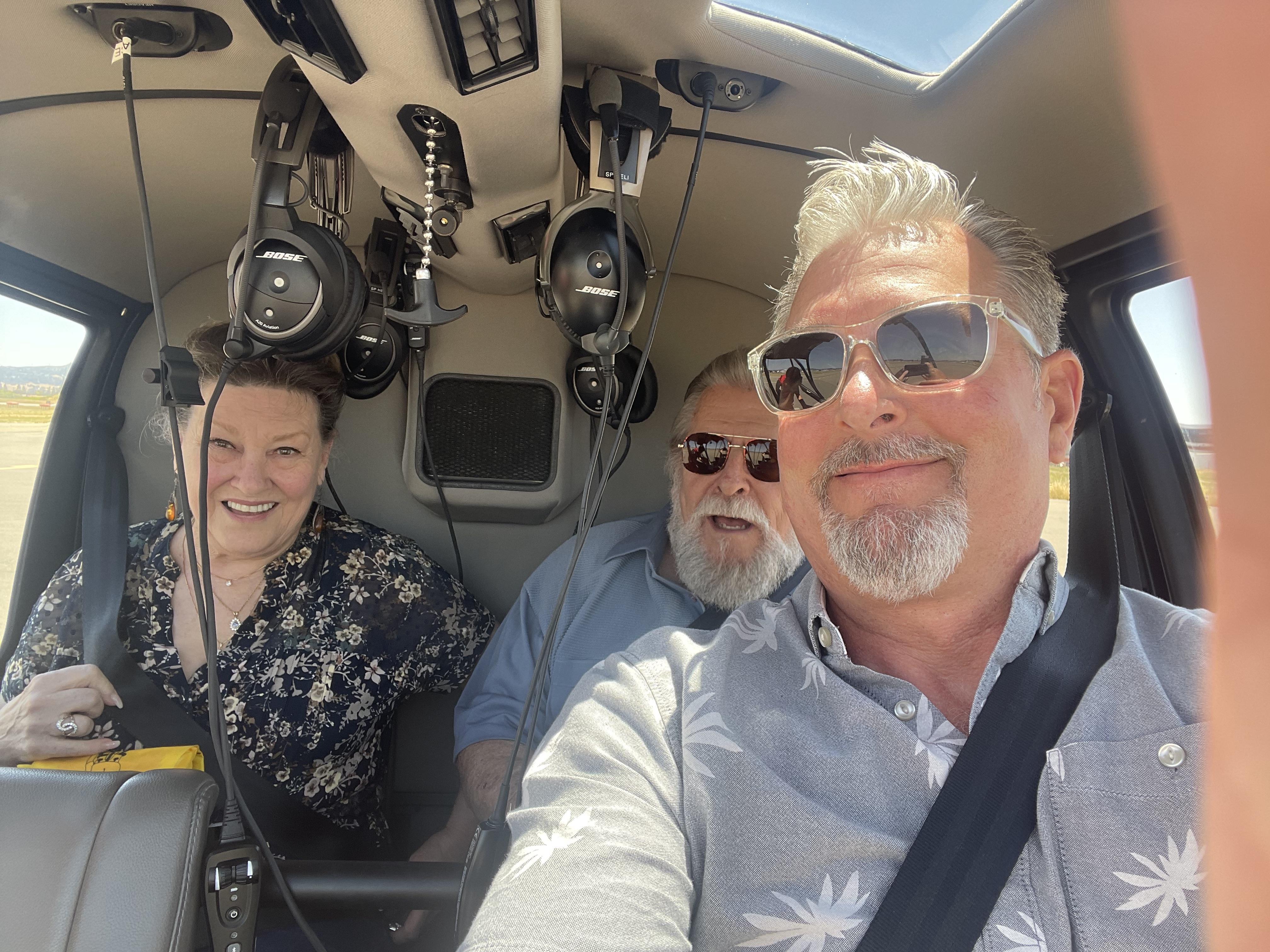 Three people enjoy a helicopter ride, wearing headsets, smiling and taking in the views.