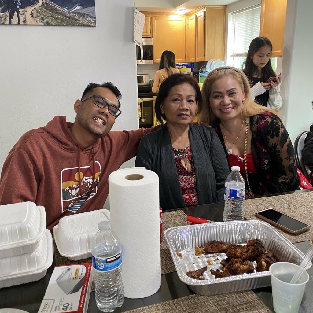 Three people sit together at a table with food, enjoying each other's company in a home.
