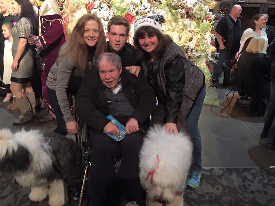 Four people pose together in front of a decorated Christmas tree with two fluffy dogs.