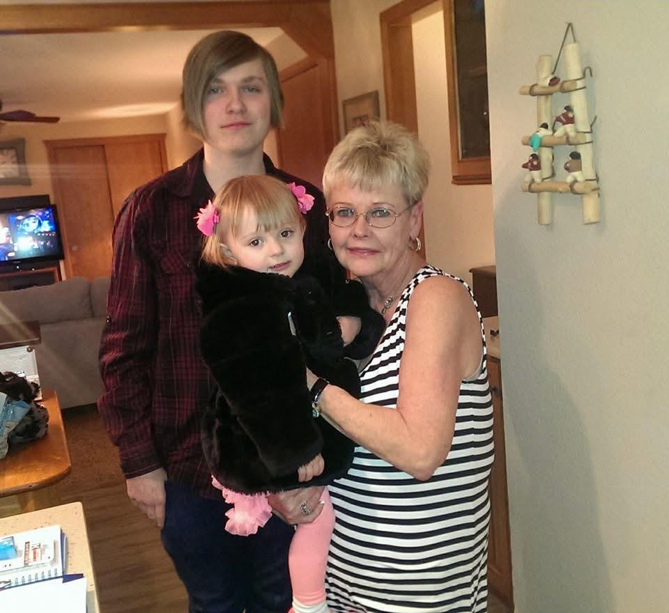 A young boy stands next to a woman and a toddler in a living room. They are in casual clothes.