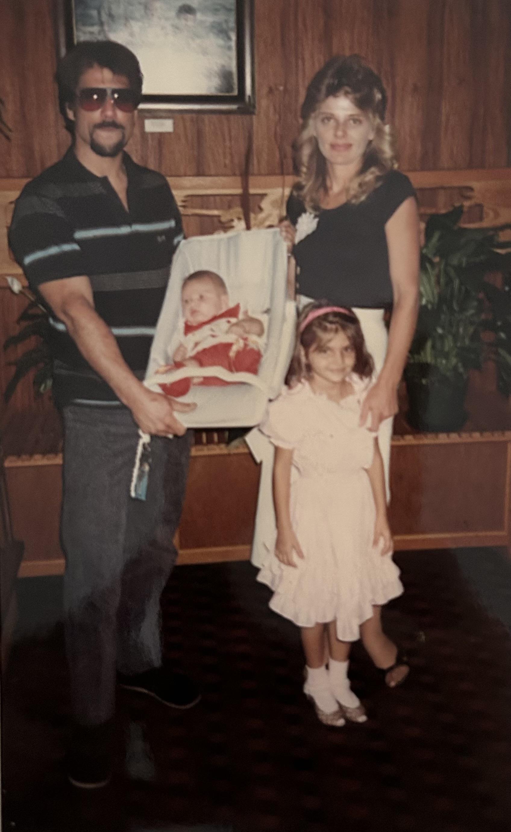 Family stands together holding a baby while an older sister looks on. They are indoors.