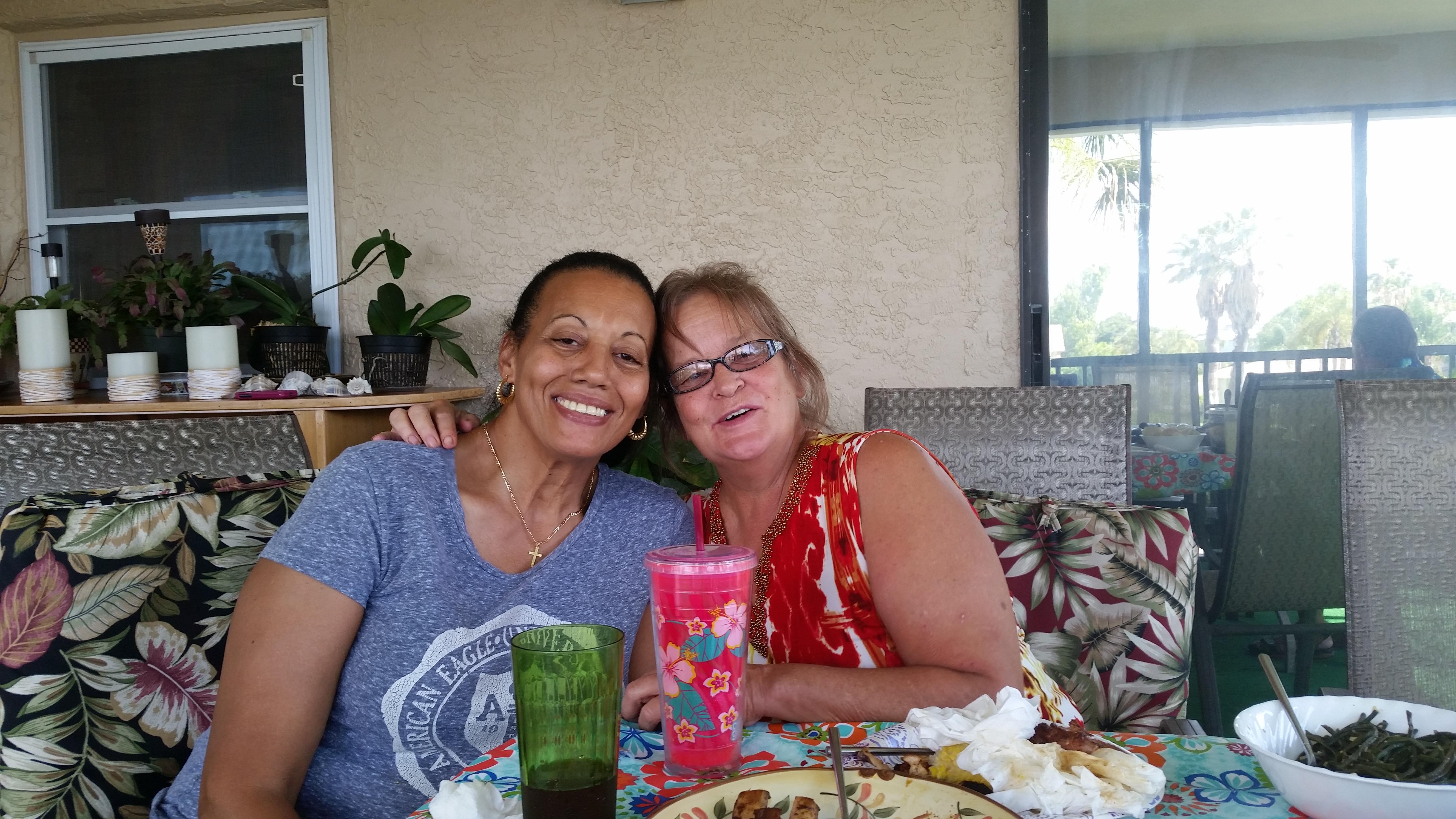 Two friends smile together while sharing a meal and drinks at a table outside during the day.