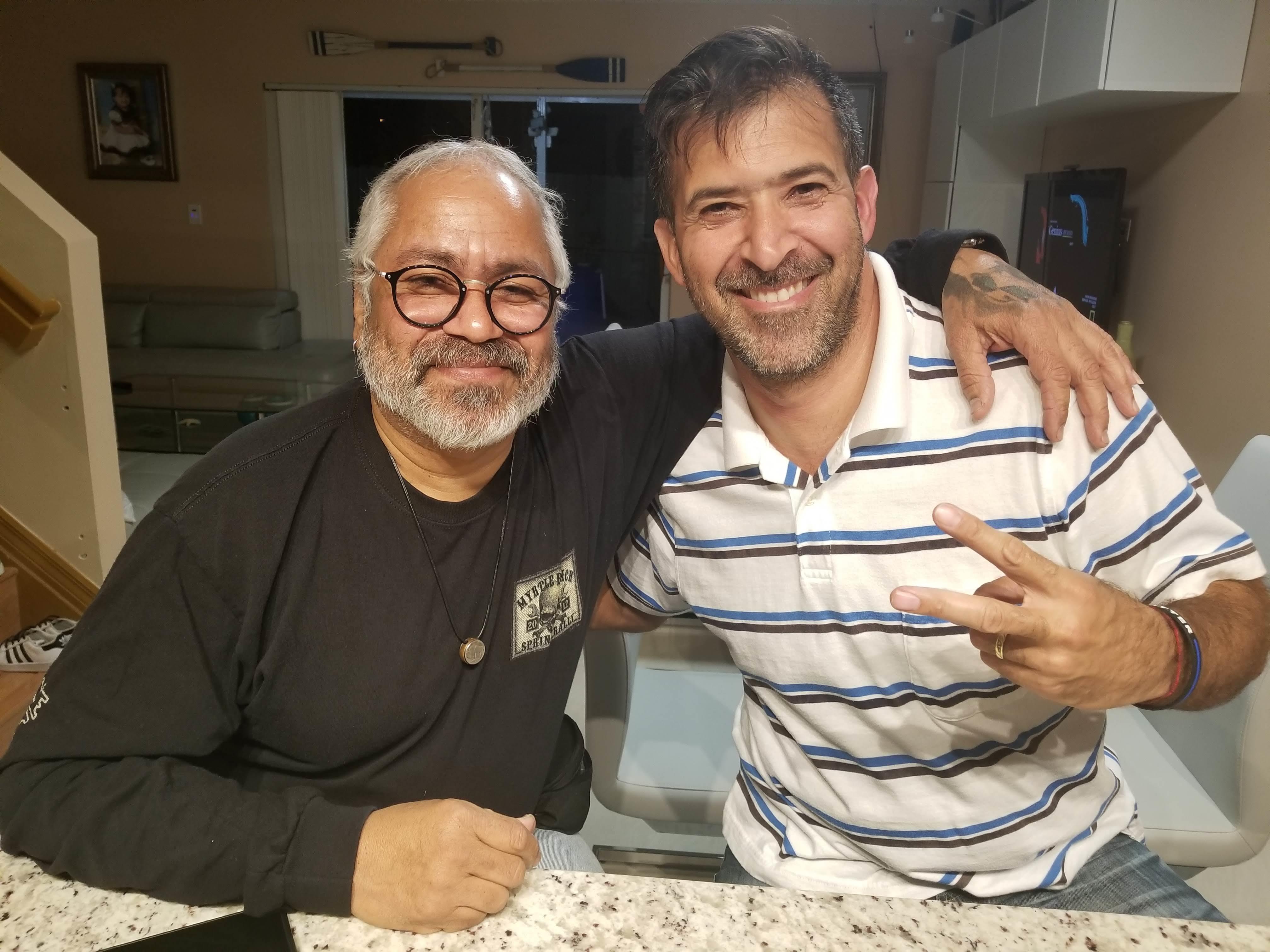 Two men are standing close together in a kitchen, both smiling and having a fun time.
