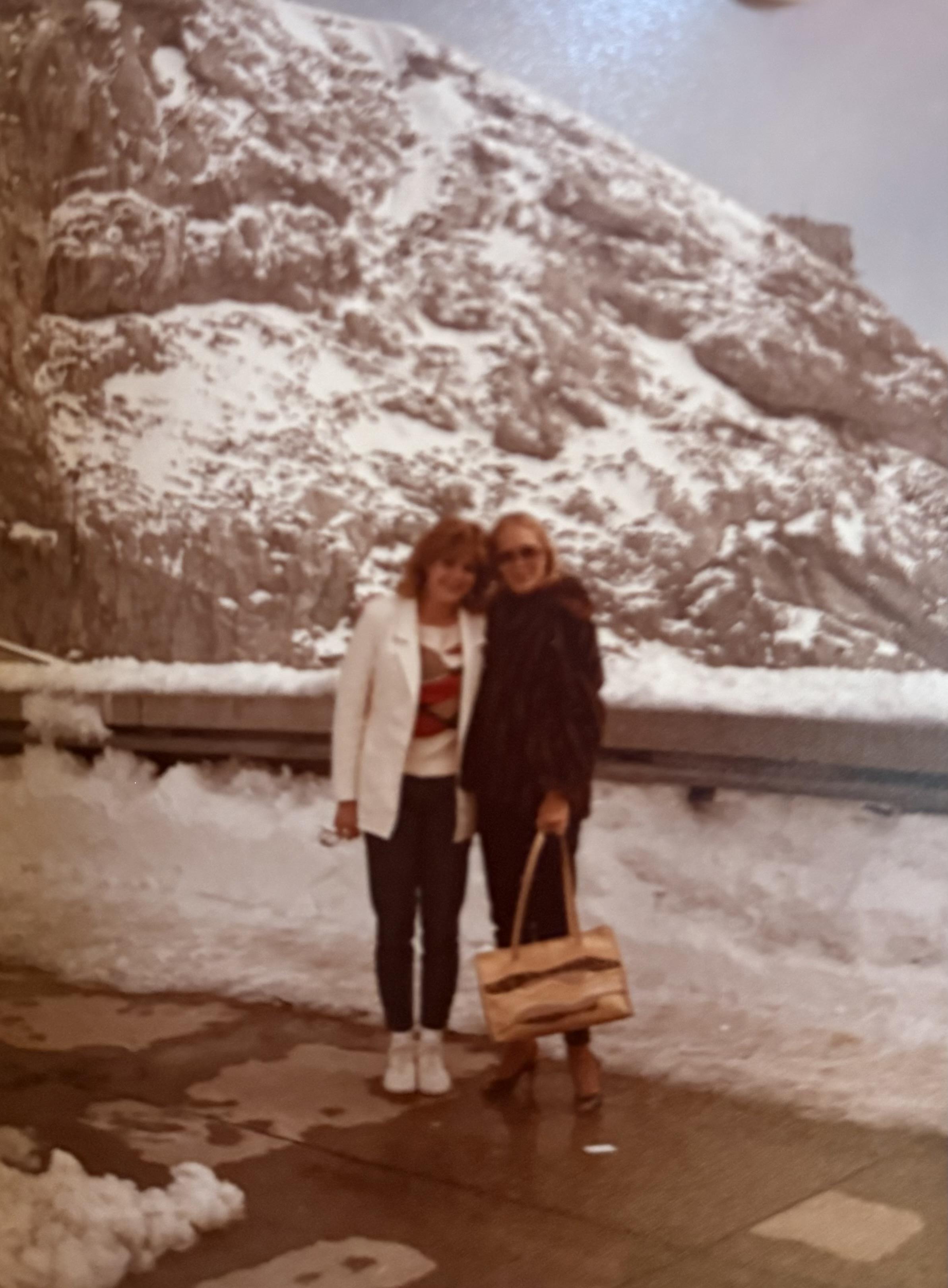Two women pose near the water with a snow-covered mountain behind them on a clear day.