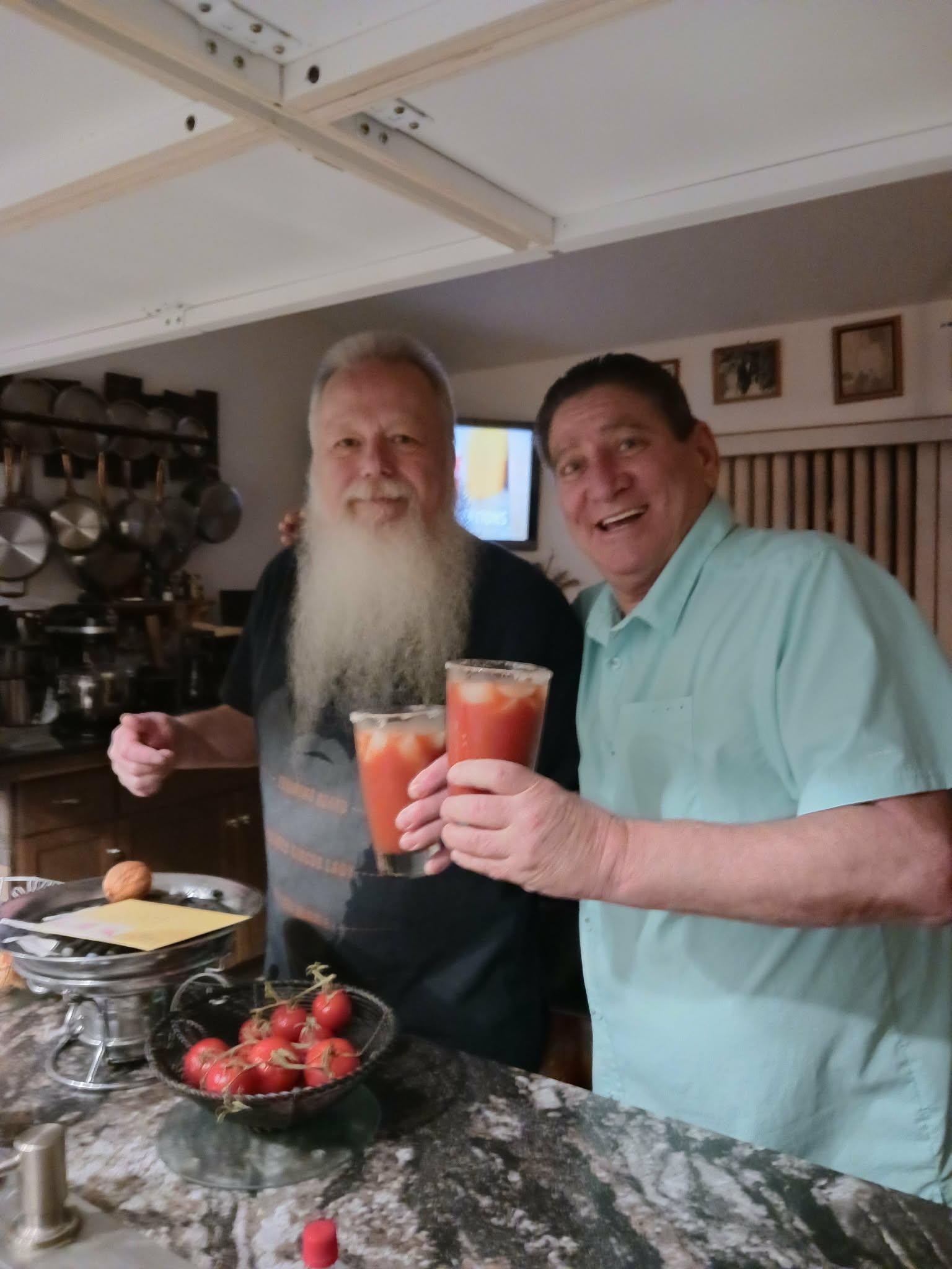 Two men enjoy drinks in a kitchen while preparing a meal with fresh tomatoes.