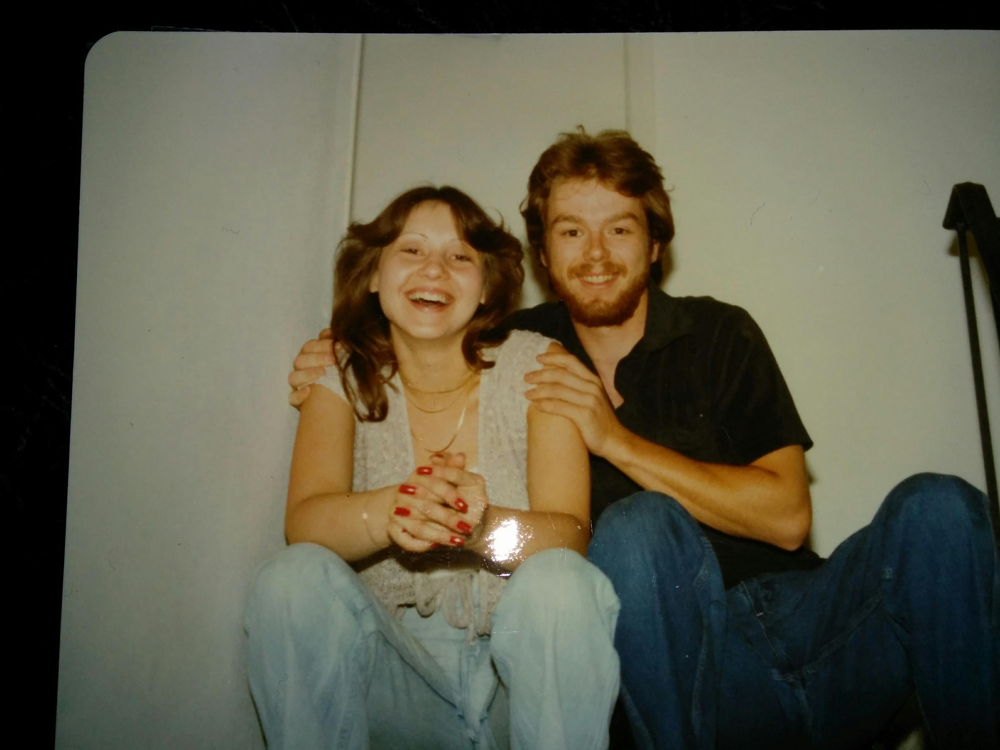 Two people sit on the floor, smiling and holding hands while sitting close together.