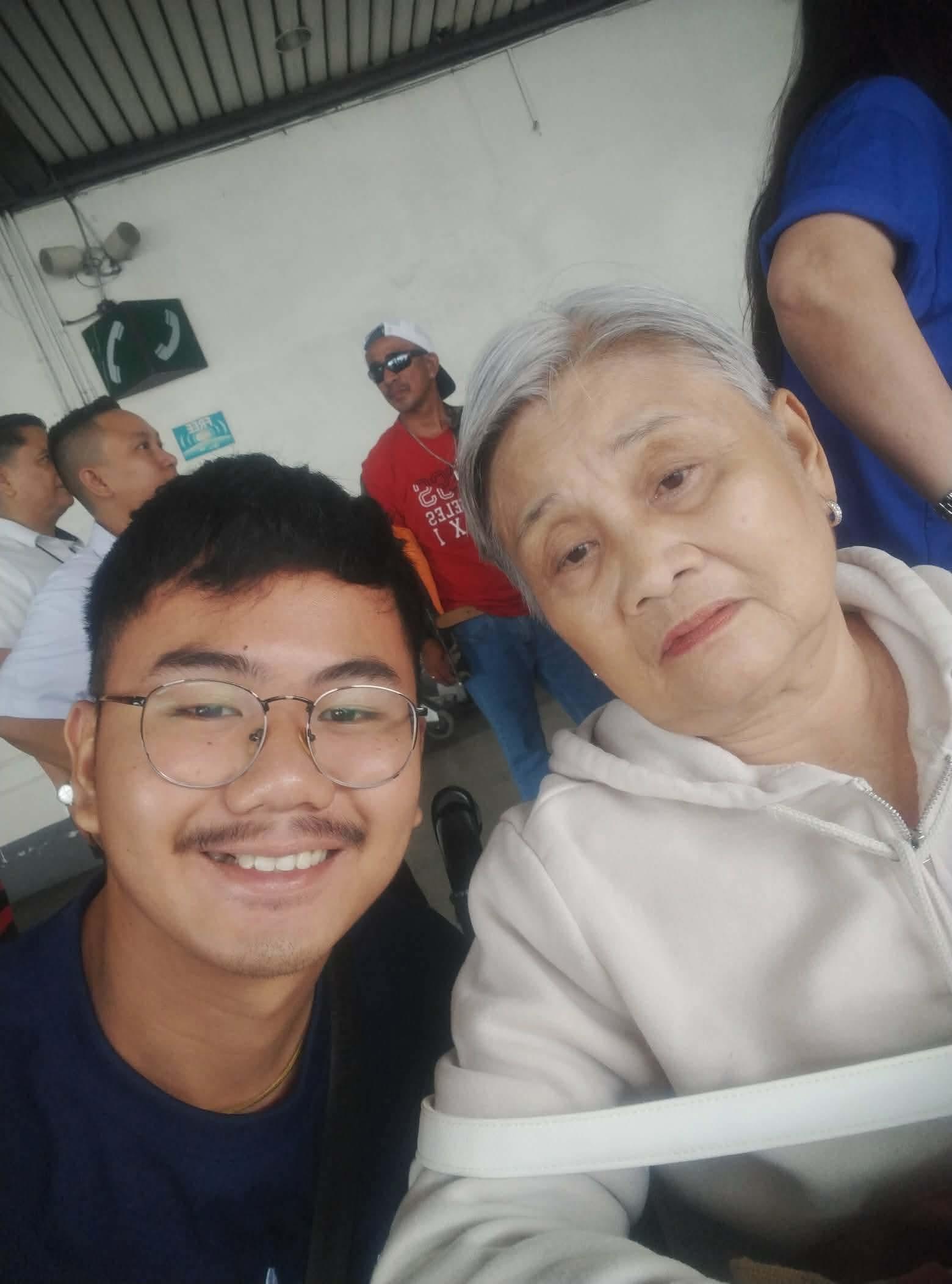 A young man smiles while sitting next to a senior woman in a busy area with people around.