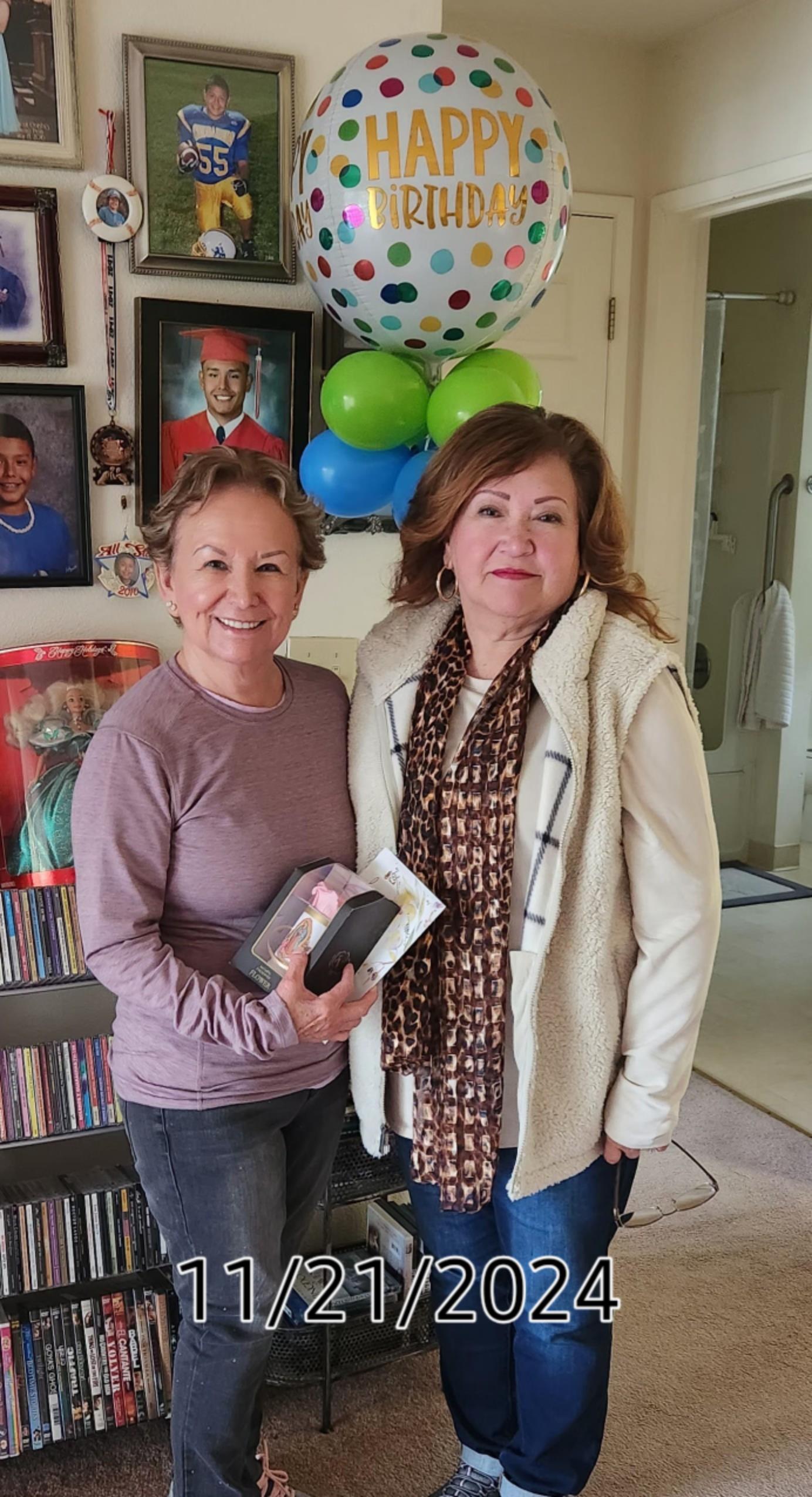 Two women smile as they hold gifts in a room with green balloons and family photos on the wall.
