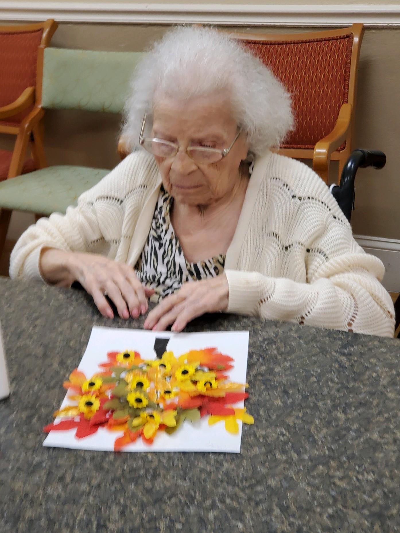 A woman makes a craft using paper flowers at a table in a senior center. She enjoys the activity.