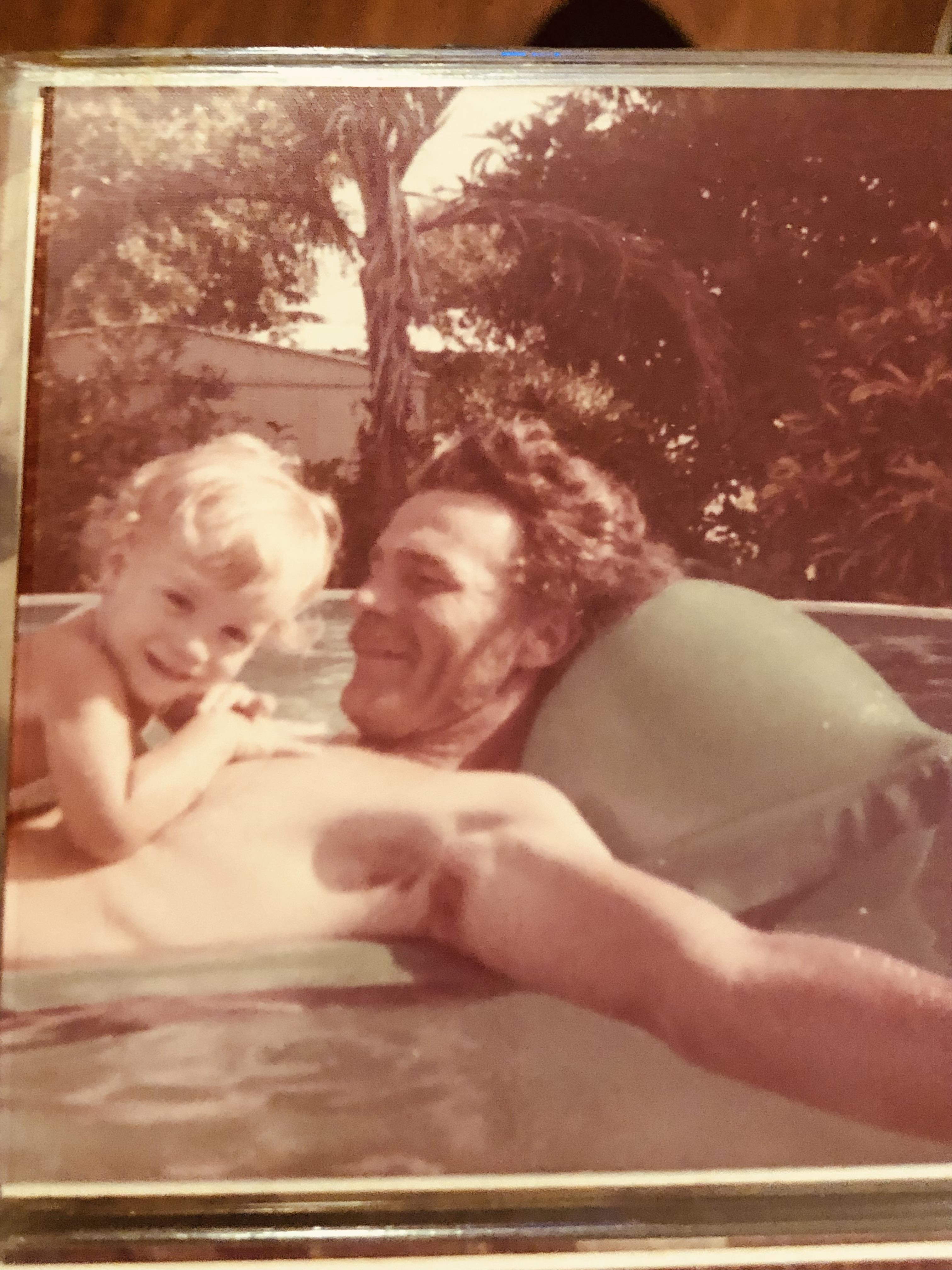 A man and a small child play in a swimming pool on a sunny day in a backyard setting.