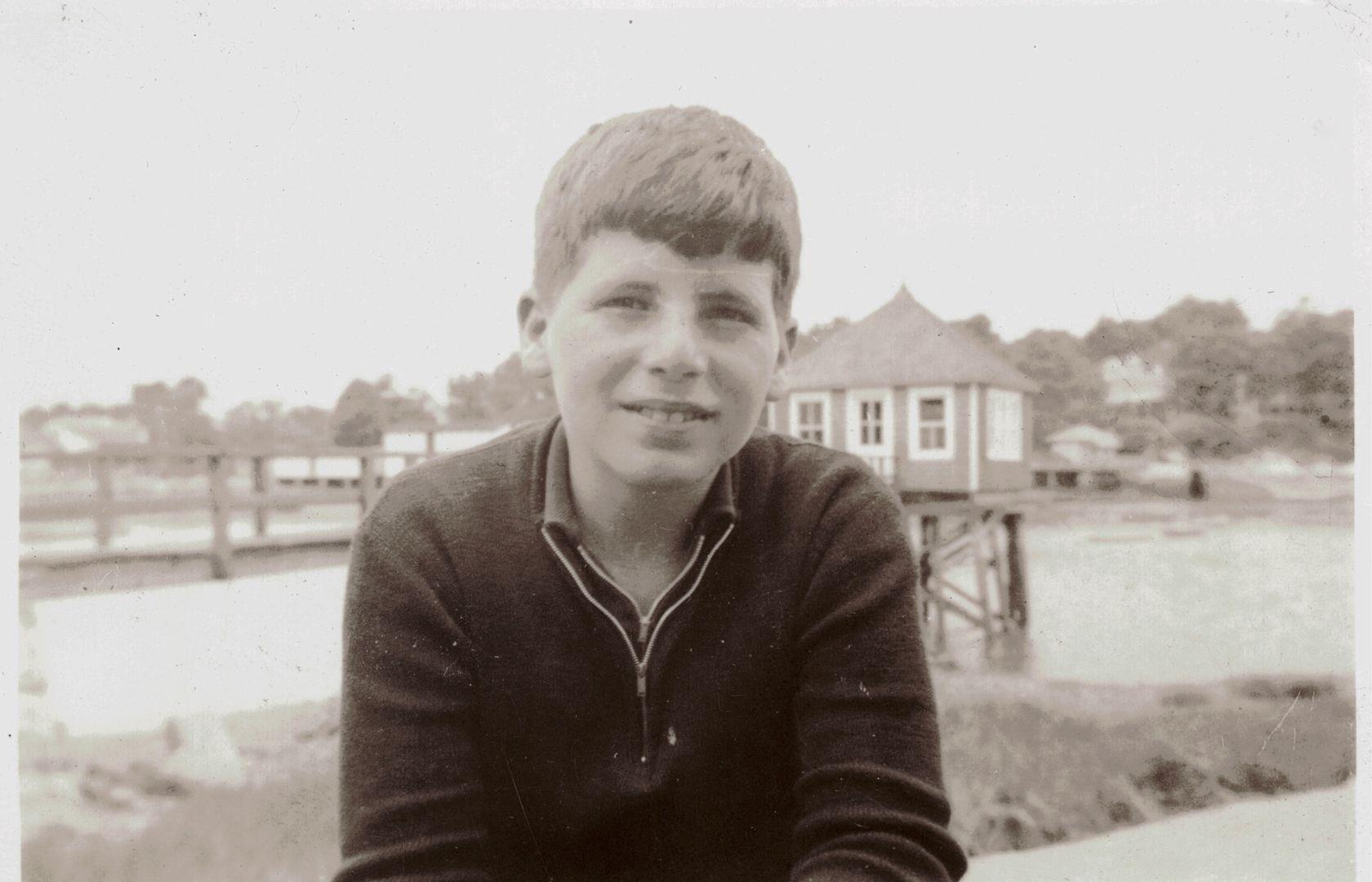 A boy is sitting by the water, looking at the camera with a smile while enjoying the day.