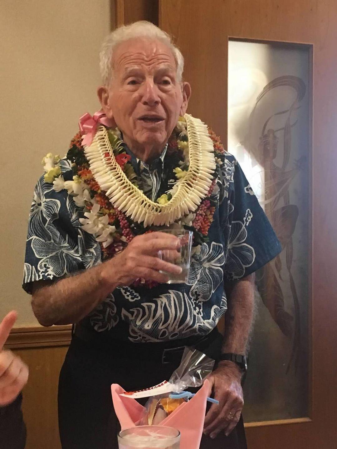 An elderly man stands with a drink in his hand surrounded by people at a social event.