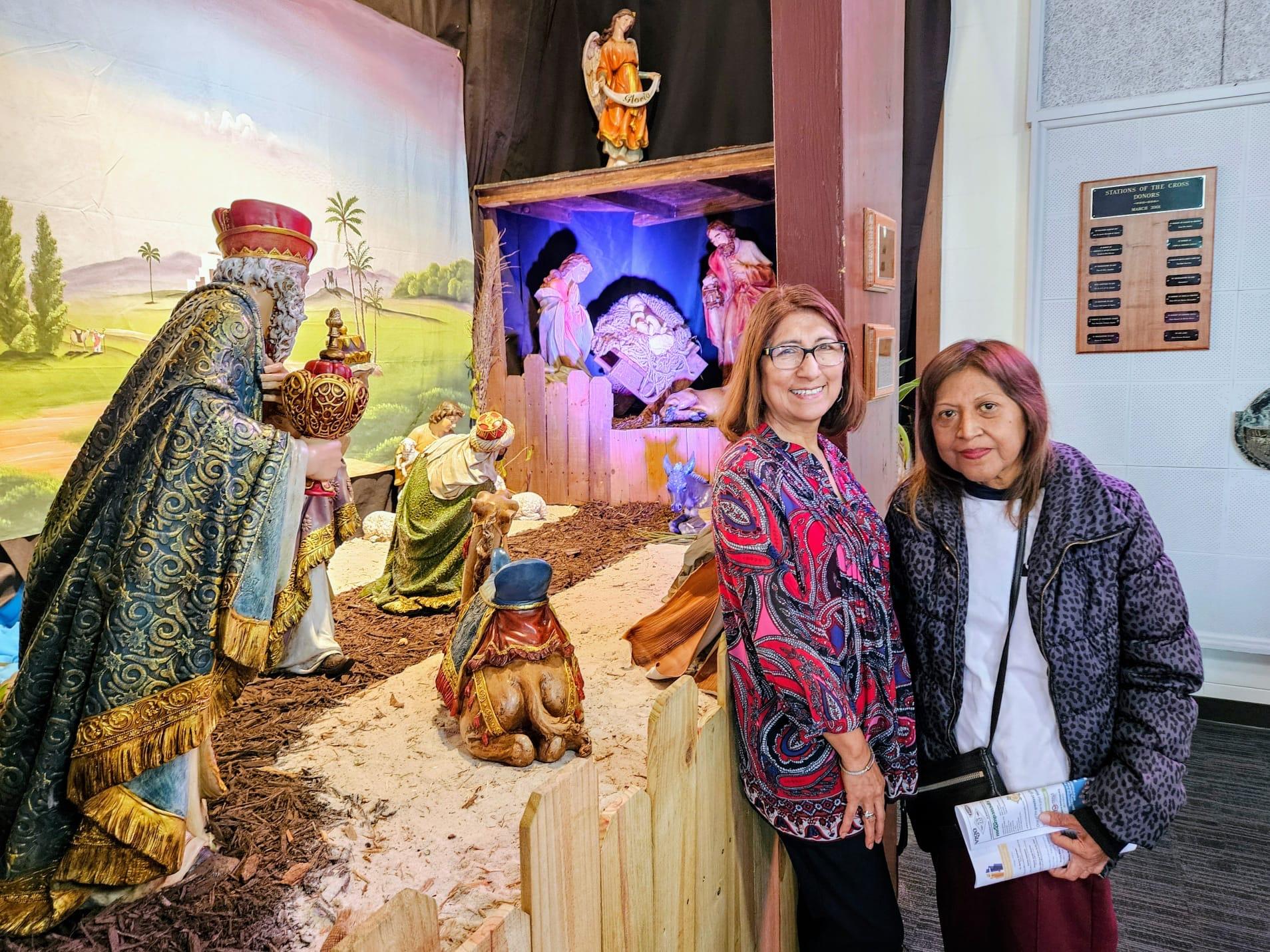 Two women stand next to a large nativity scene decorated for a holiday event in a public space.