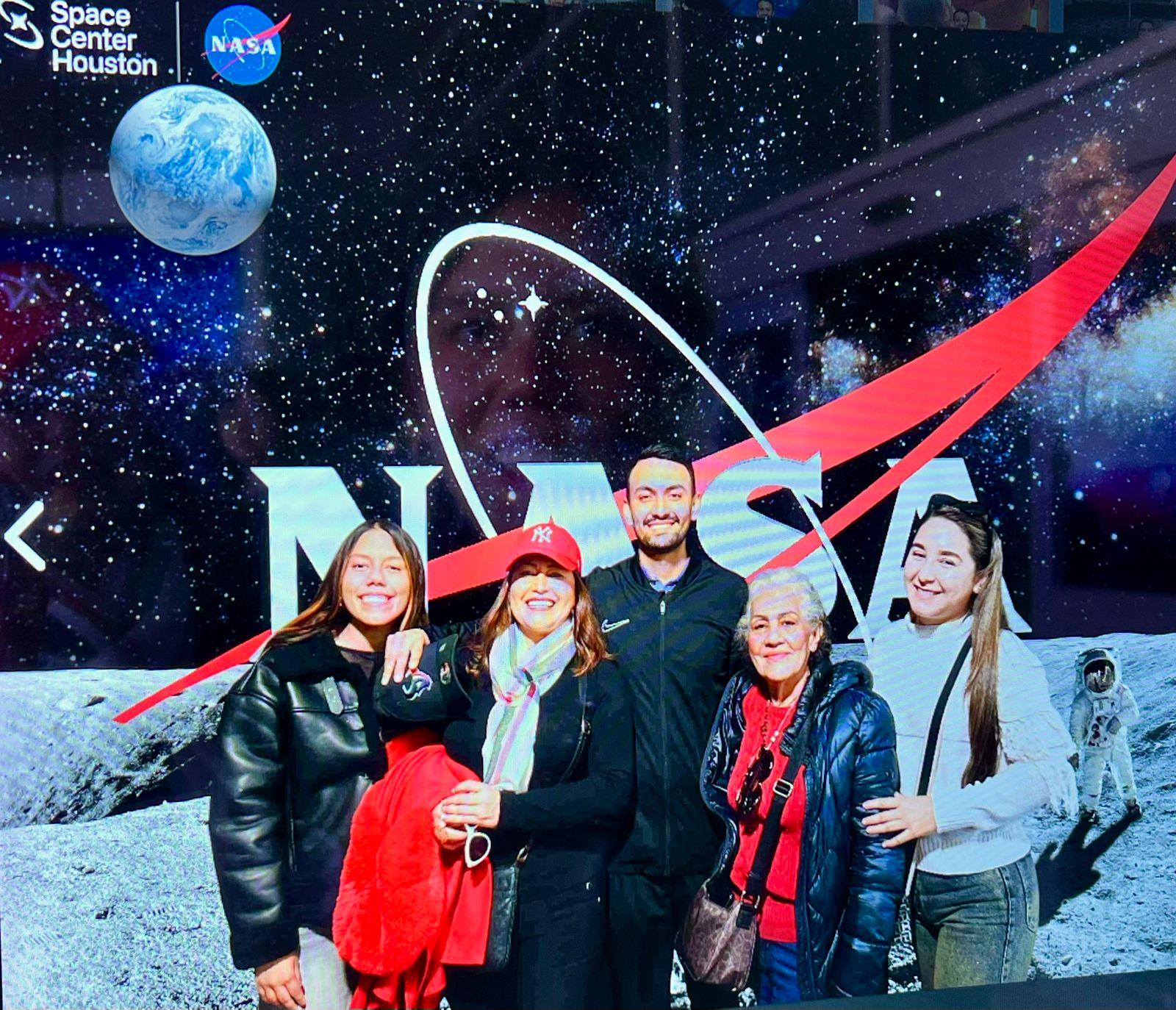 A group of six people poses happily in front of a NASA sign at a space exhibit. They smile together.