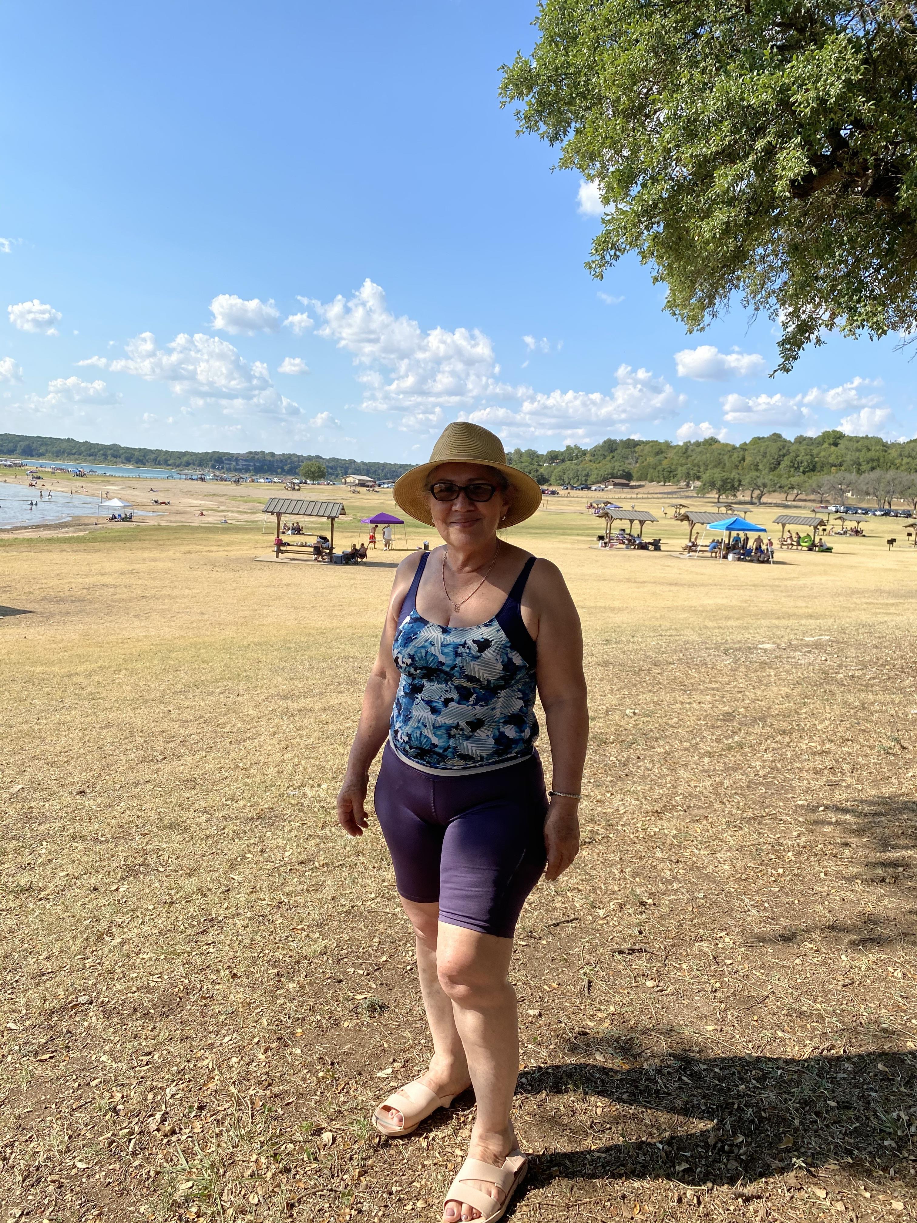 A woman smiles by the lake on a sunny day, wearing a hat, with others in the background.