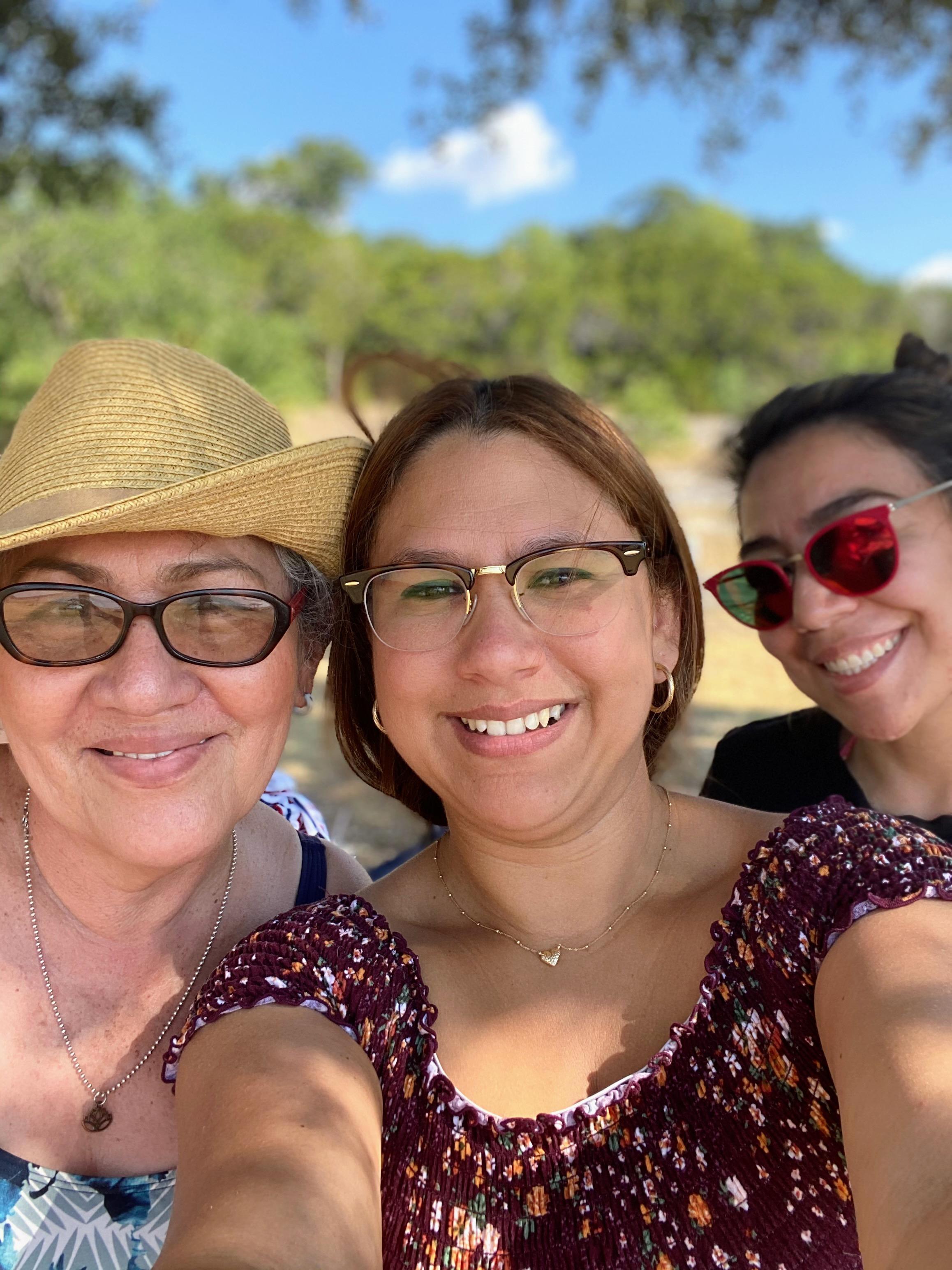 Three women take a selfie while enjoying time at a park with trees and water during the day.
