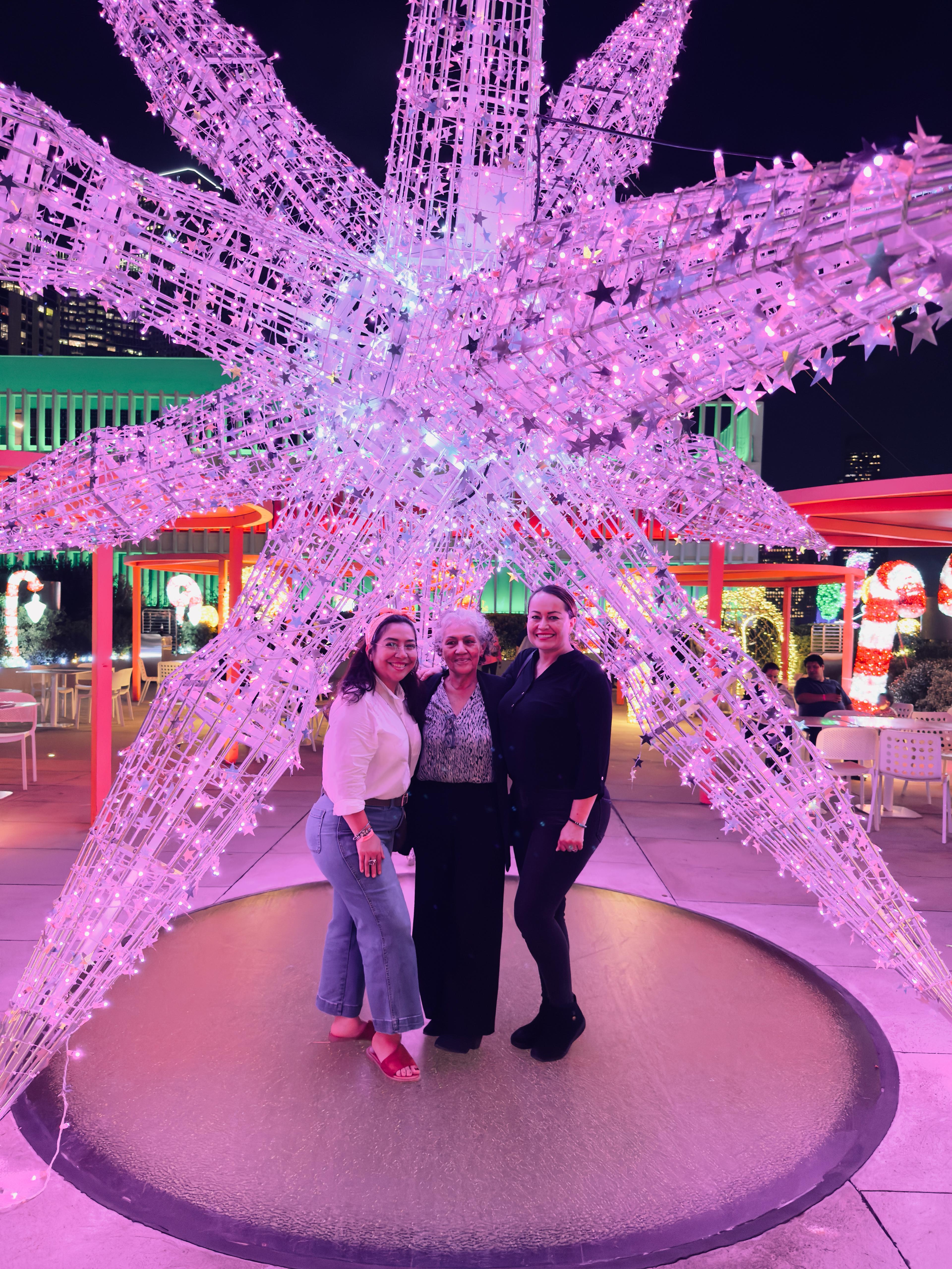 Three women smile together in front of an illuminated star decoration during a festive gathering.