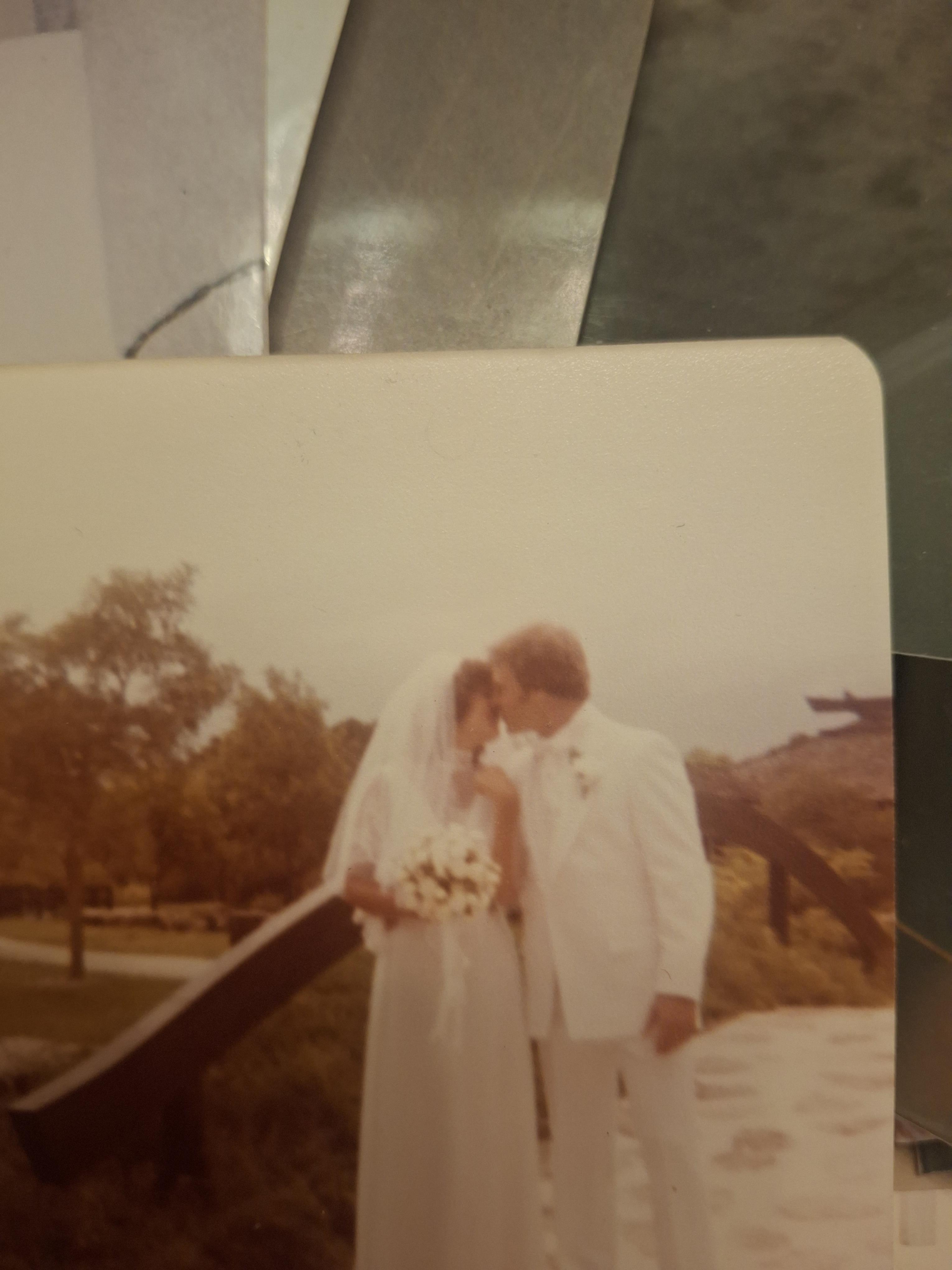 A couple stands close together, kissing in a garden. They wear wedding clothes and embrace.