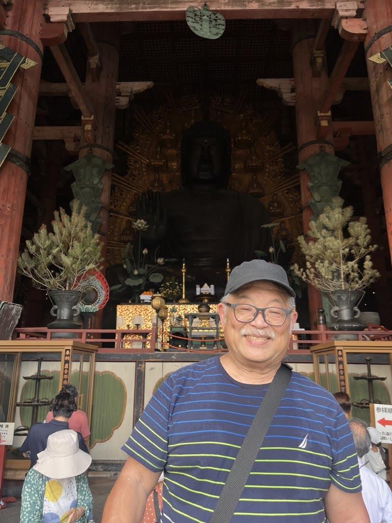 A man stands in a temple with a large statue behind him and decorations around.