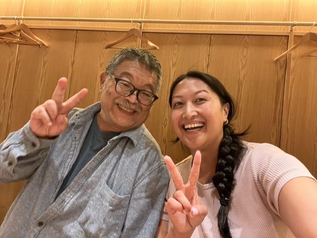 Two people are smiling and making peace signs while sitting together in a bright room.
