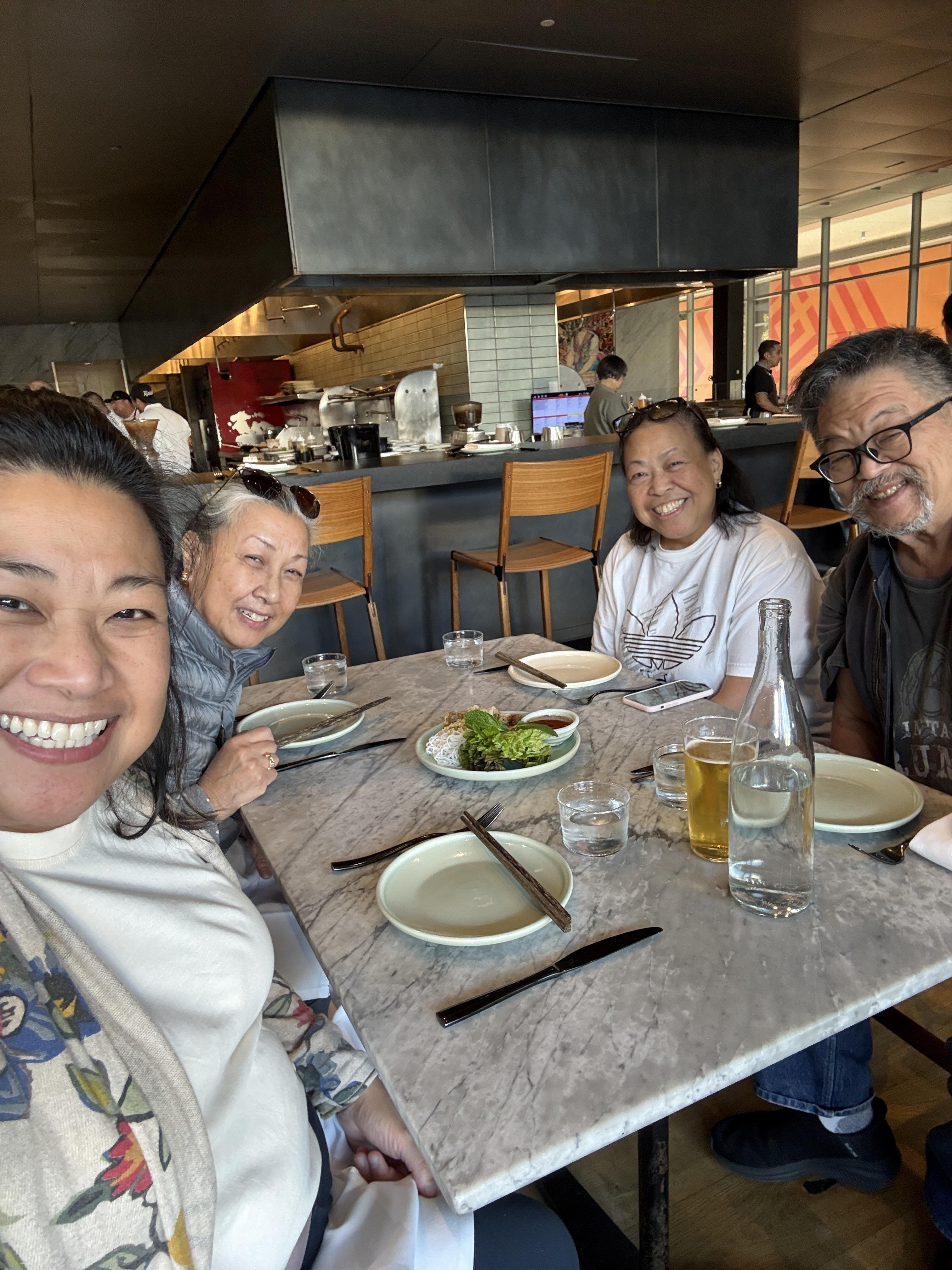 Four friends smile while sitting at a table ready to eat in a busy restaurant setting.