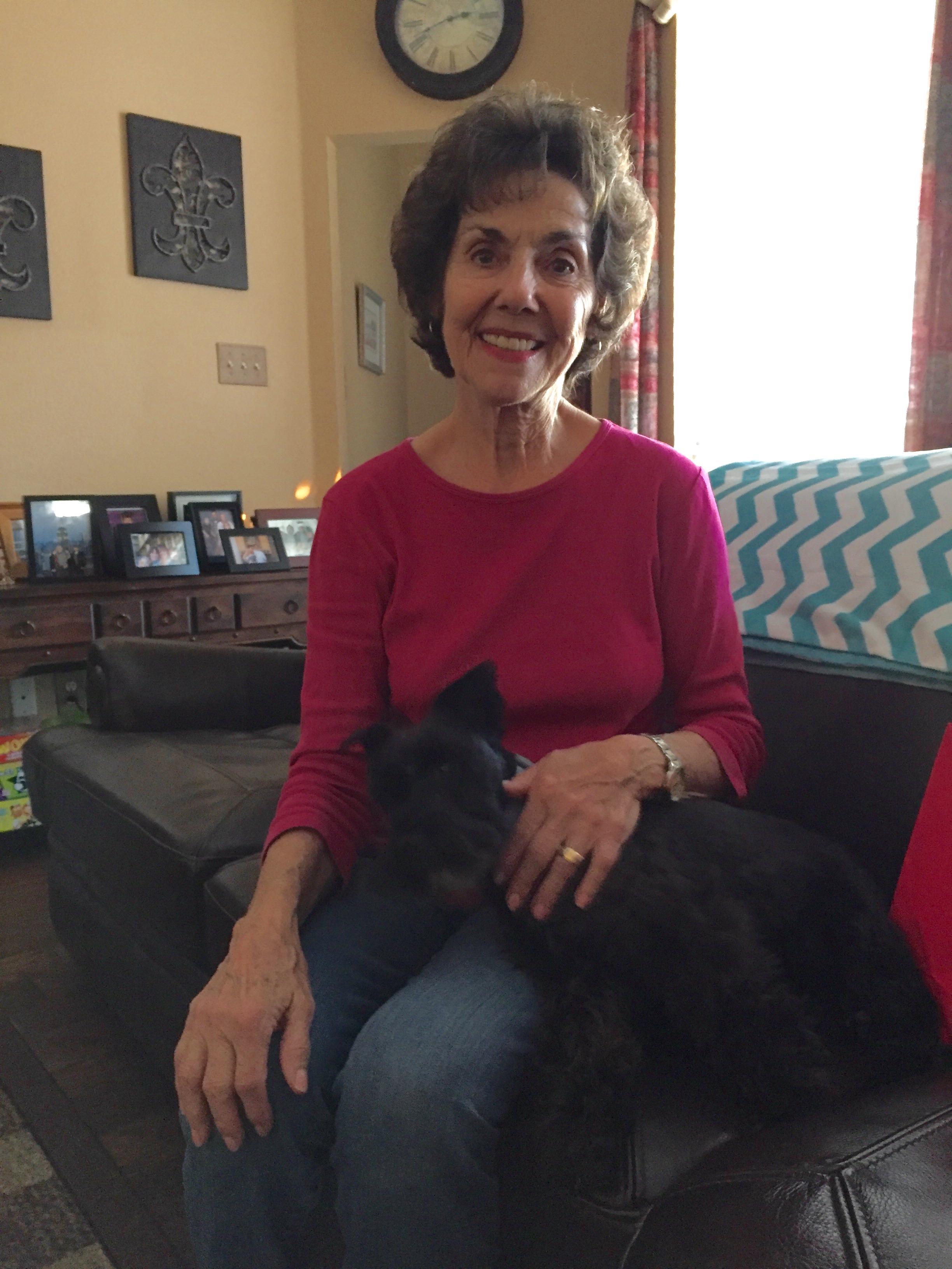 A woman enjoys time indoors with a dog resting on her lap and family photos in the background.